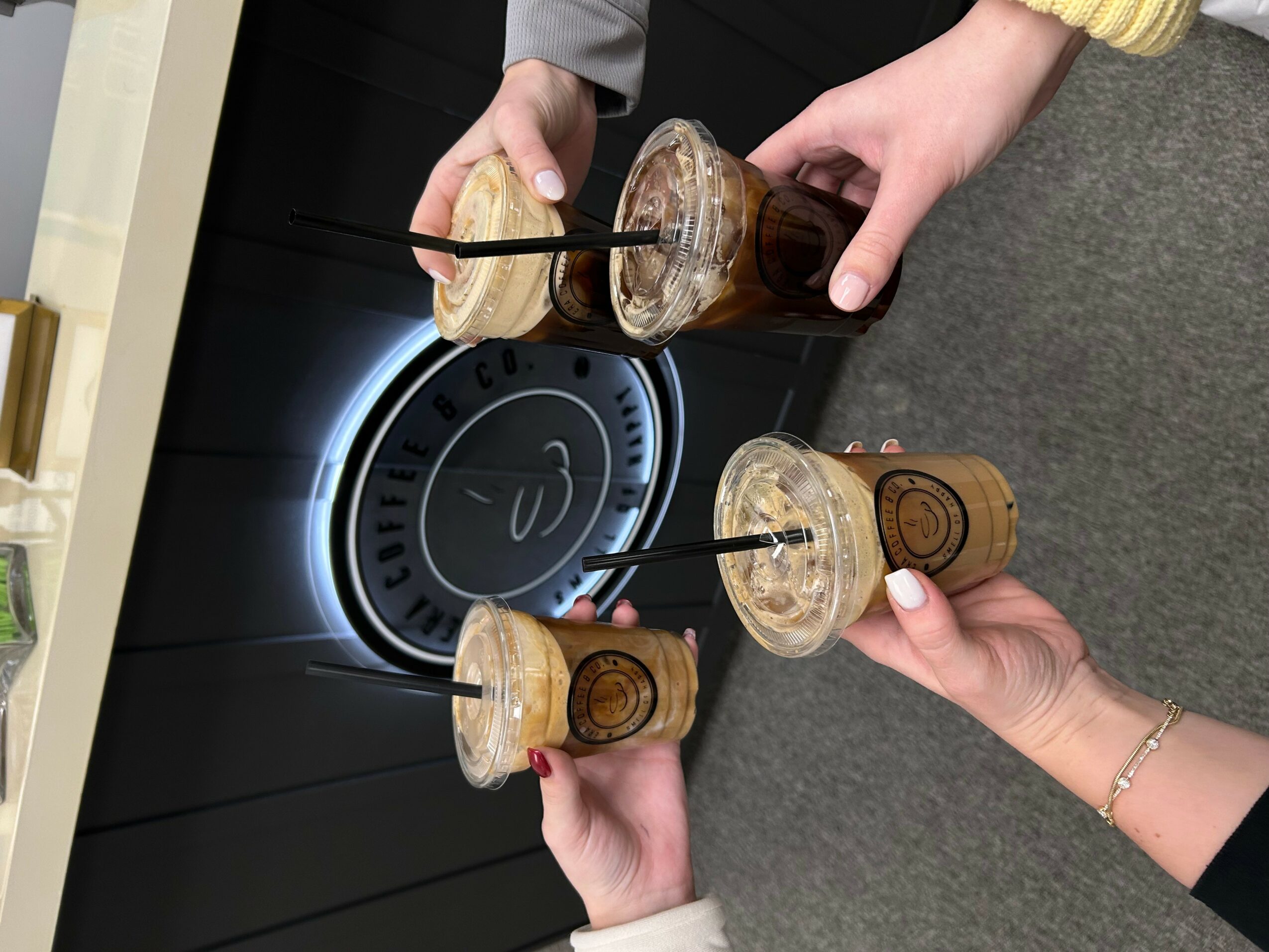 Three people hold drinks with straws, clinking glasses in front of a clock with a smiley face. Gray carpeted floor visible.