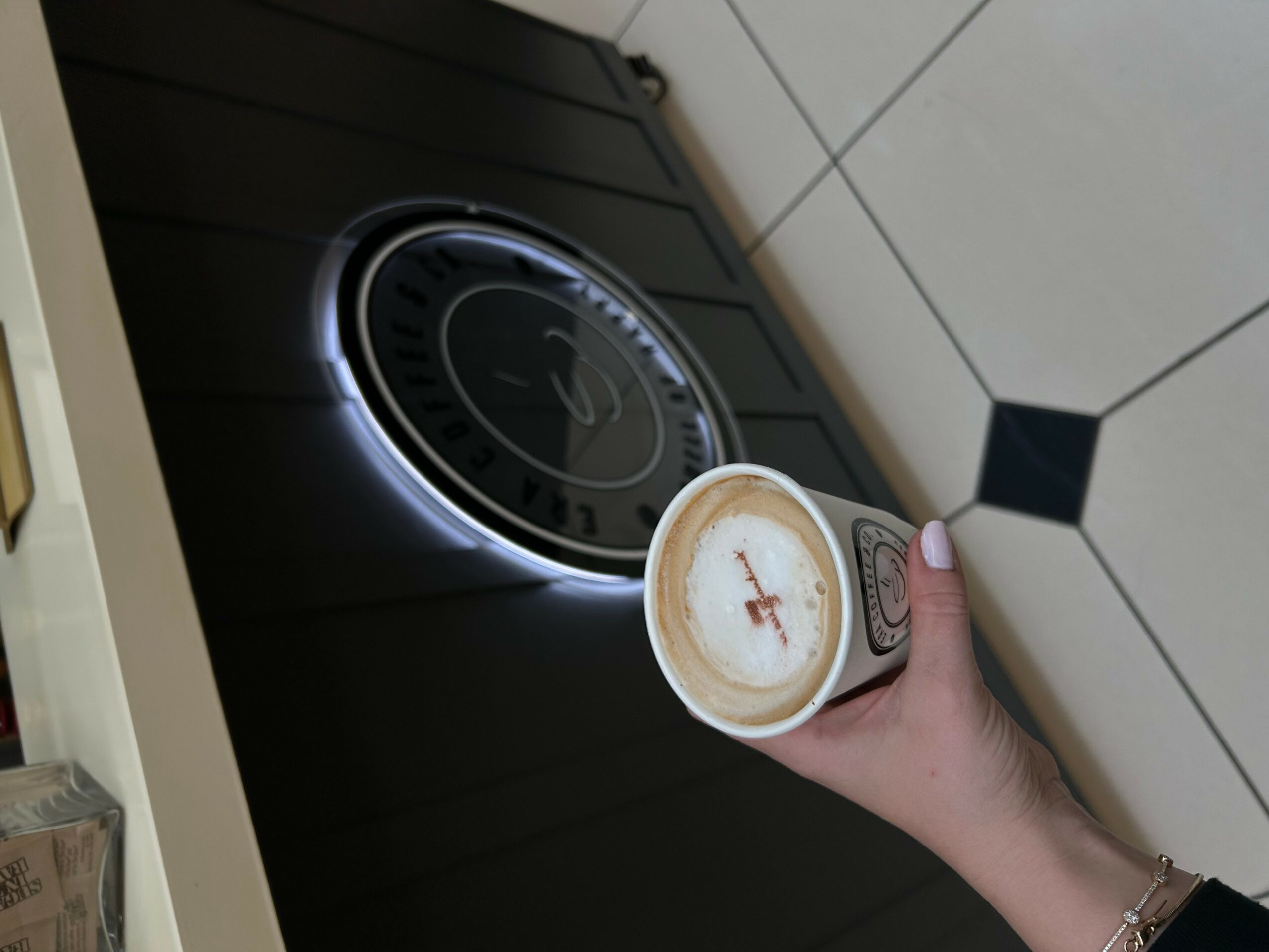Person holding a cup of coffee near a stovetop with a black surface and a circular burner, tiled wall in background.