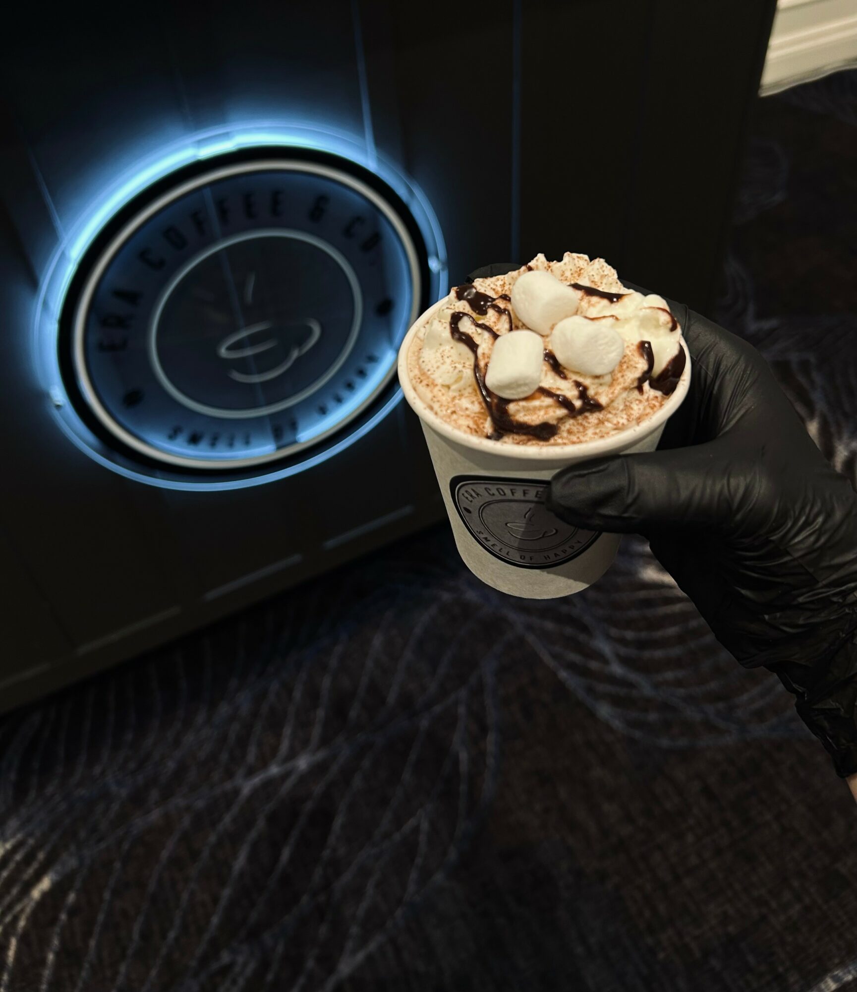 Person wearing a black glove holds a cup of ice cream with toppings near a glowing coffee machine display.