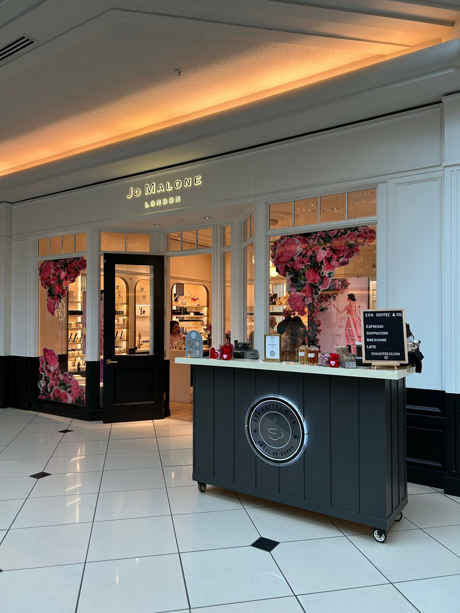 Lobby with reception desk, floral decorations, and a digital display, viewed from the side with tiled floor and ceiling lighting.
