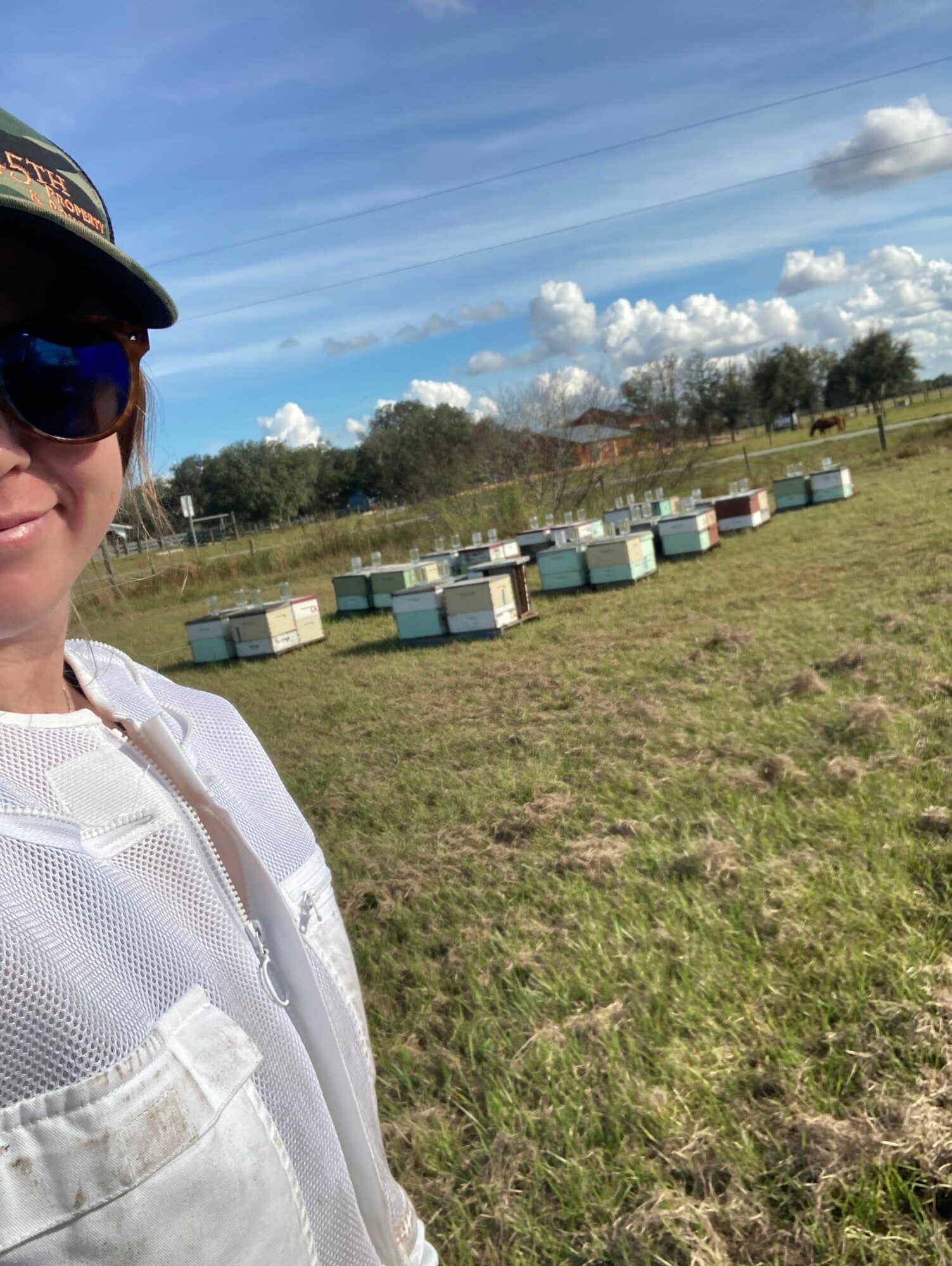 Person wearing white clothing and sunglasses outdoors with stacked containers and trees in the background.
