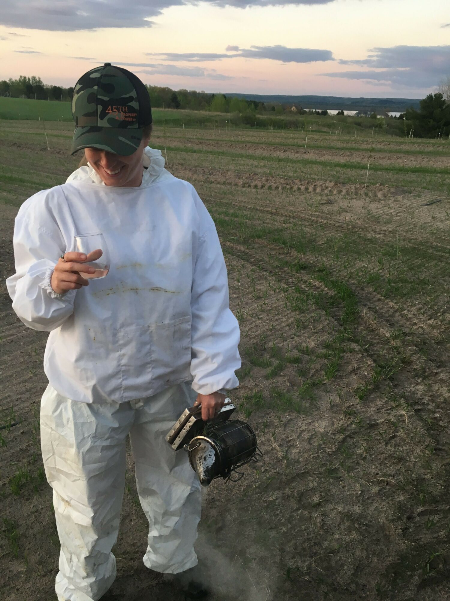 Person in white protective suit and helmet standing outdoors, holding a device, with a field and sky in the background.