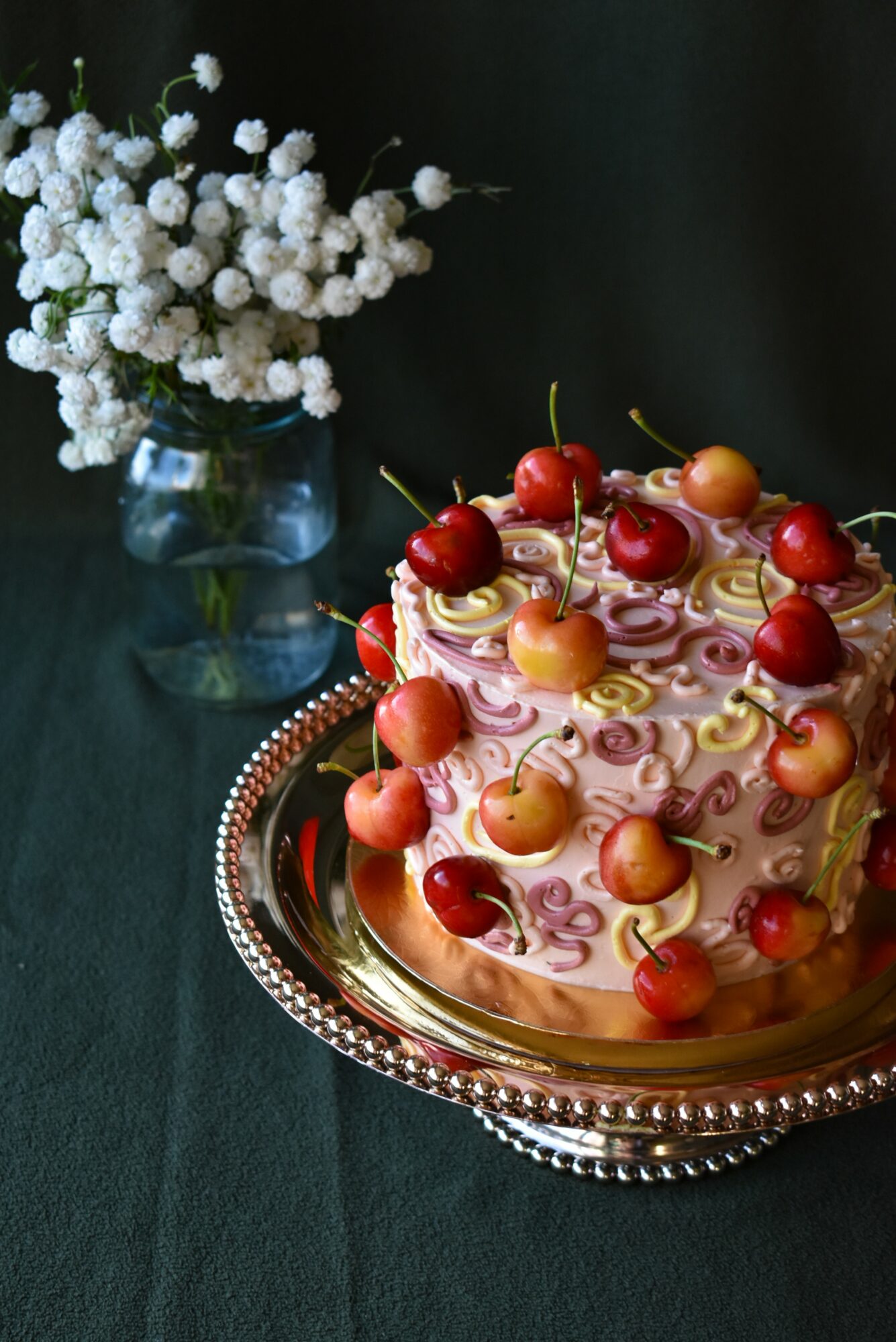 A decorated cake with cherries and cream on a silver tray, with a vase of white flowers in the background.