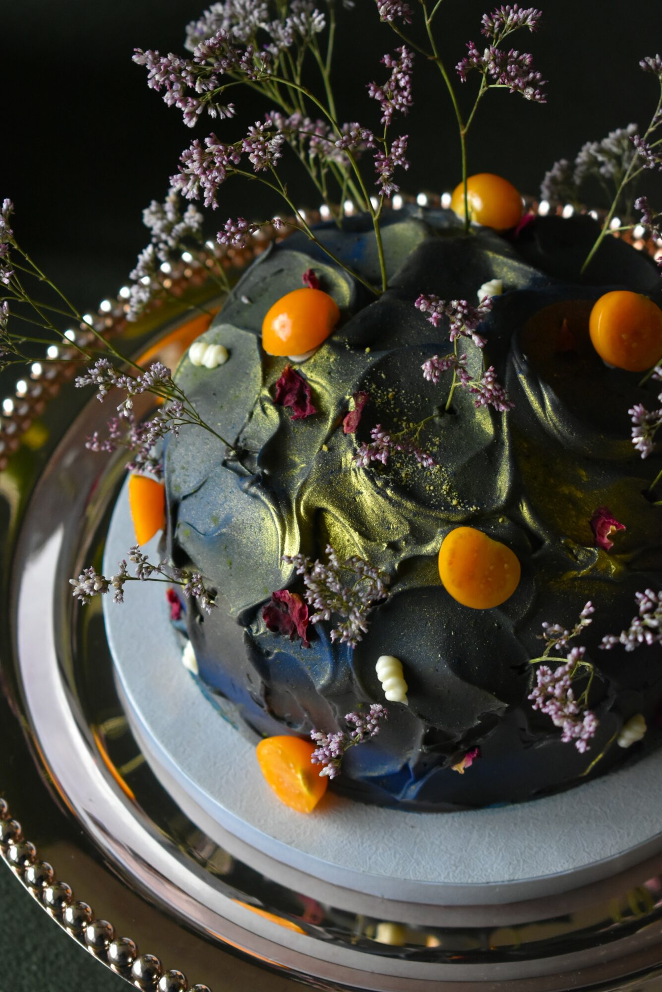 Cake decorated with black icing, orange spheres, and small purple flowers on a silver tray.