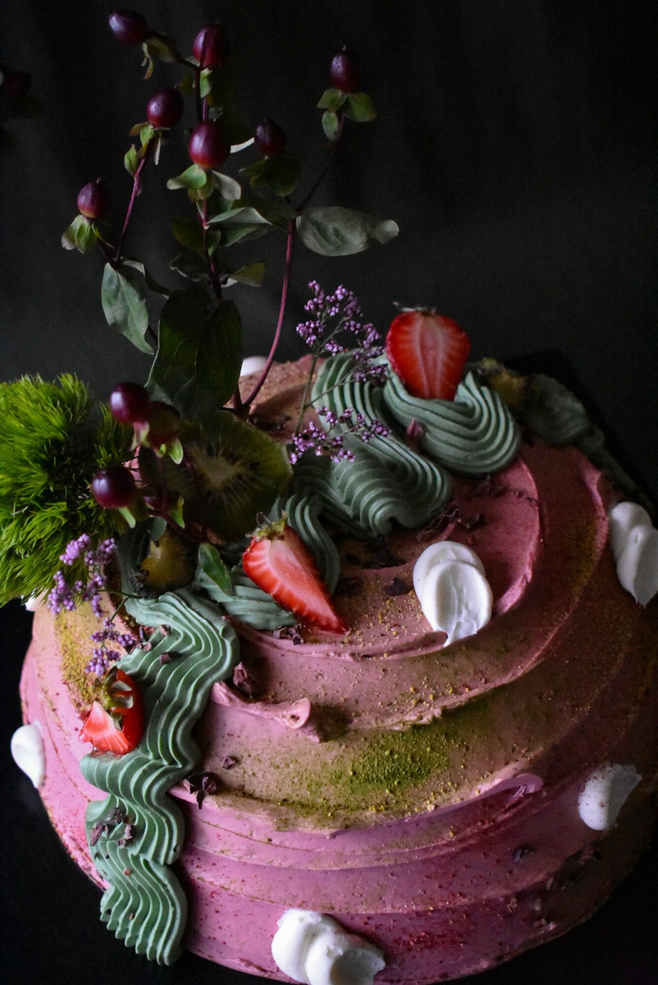 Pink frosted cake decorated with strawberries, green and purple icing, and small white meringues, against a dark background.