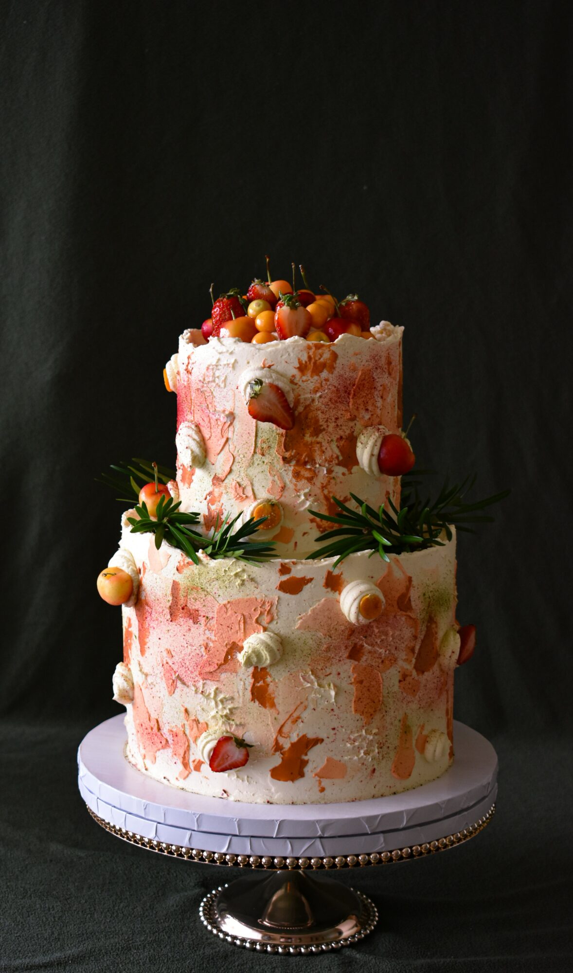 Two-tiered cake decorated with fruit and greenery, on a silver stand against a dark background.