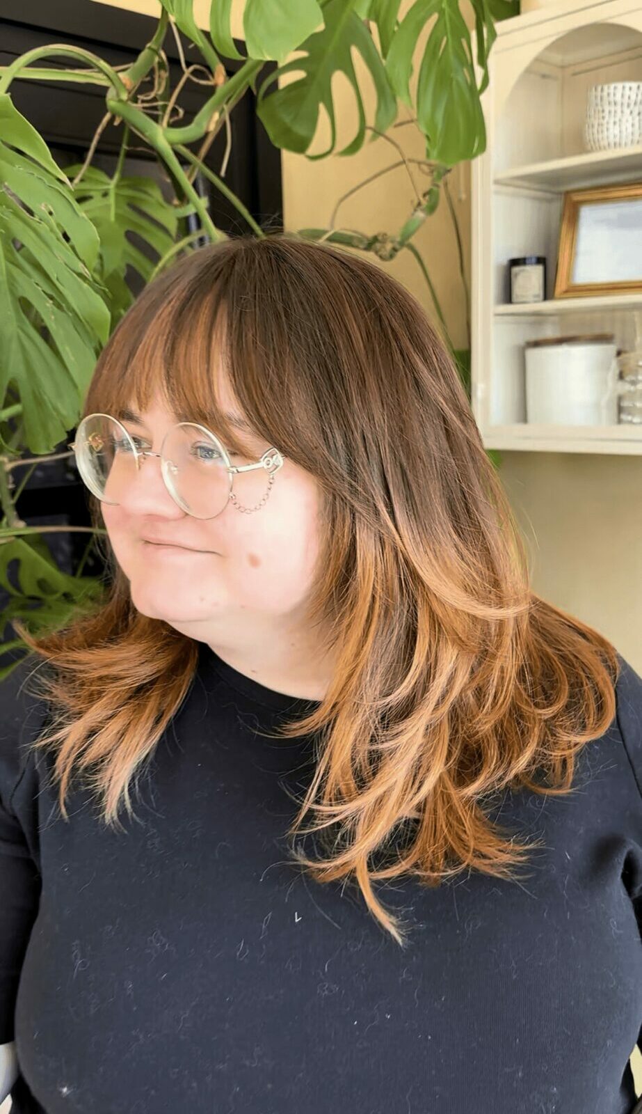 Woman with glasses and shoulder-length hair in front of green plants and a shelf, wearing a black top.
