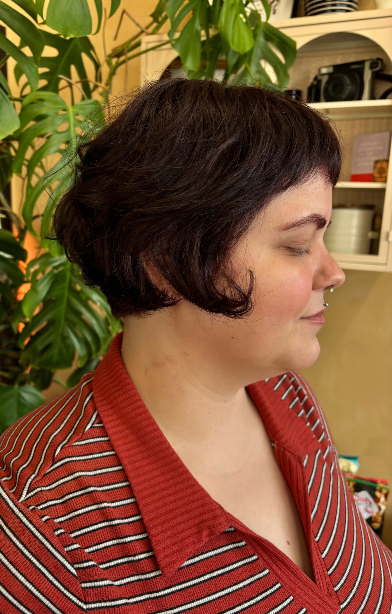 Profile of a woman with short dark hair, wearing a red and white striped shirt, standing indoors near plants and shelves.