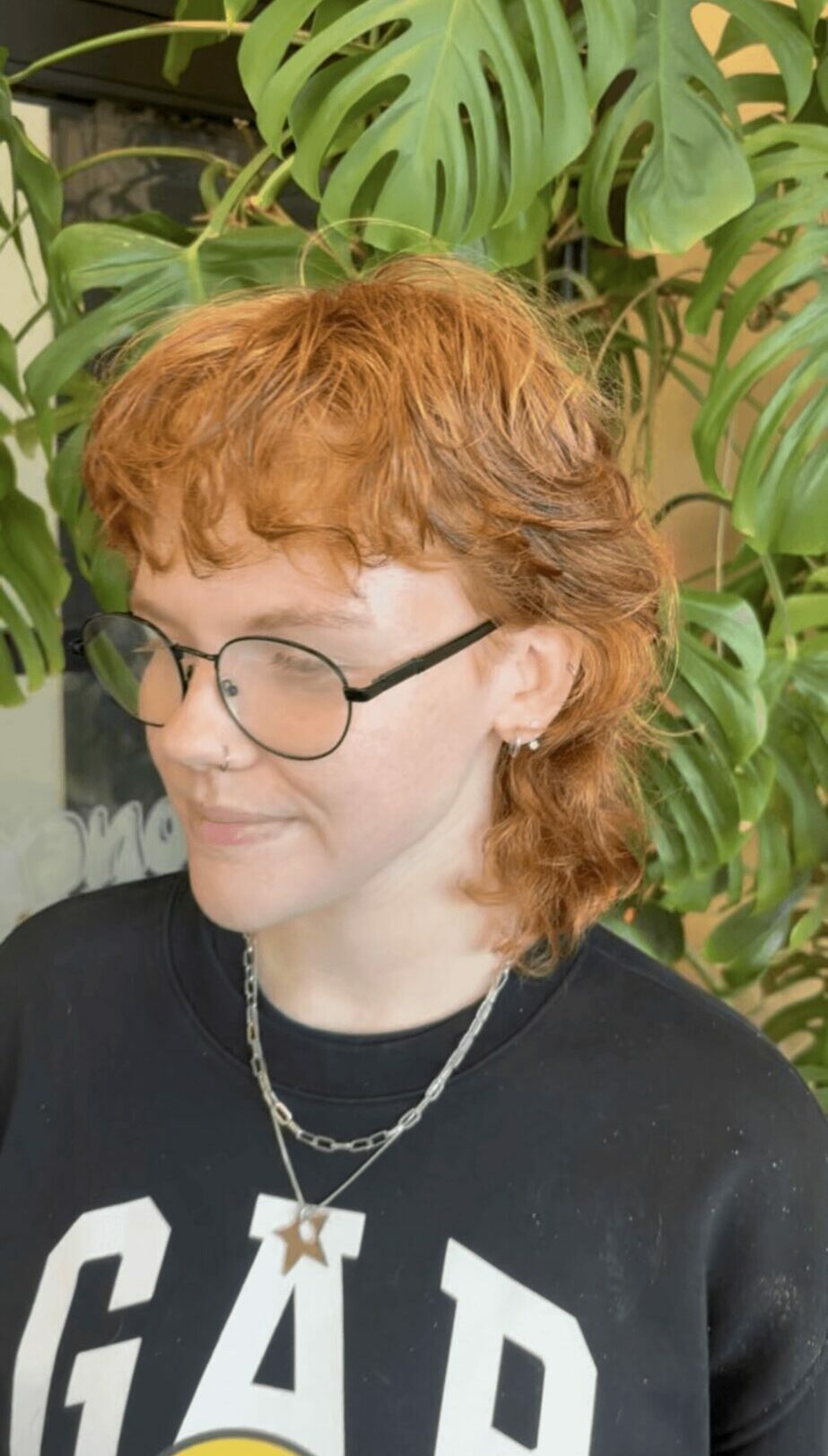 Person with curly red hair and glasses, wearing a black shirt with white letters, standing in front of green plants.