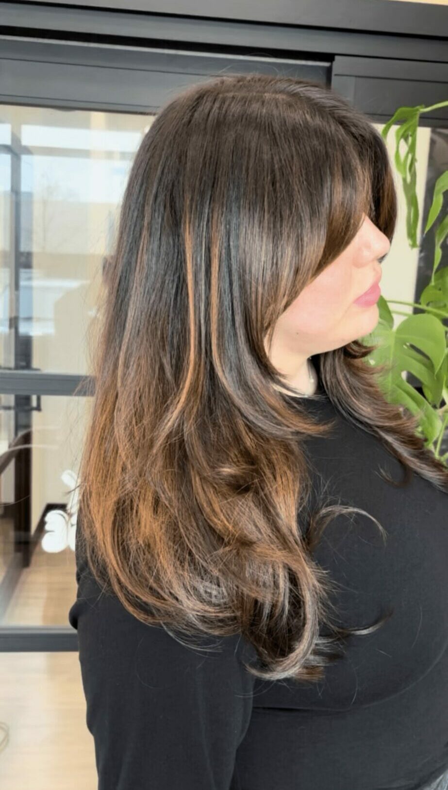 Side view of a woman with long, wavy brown hair and a black top, standing indoors near a plant.