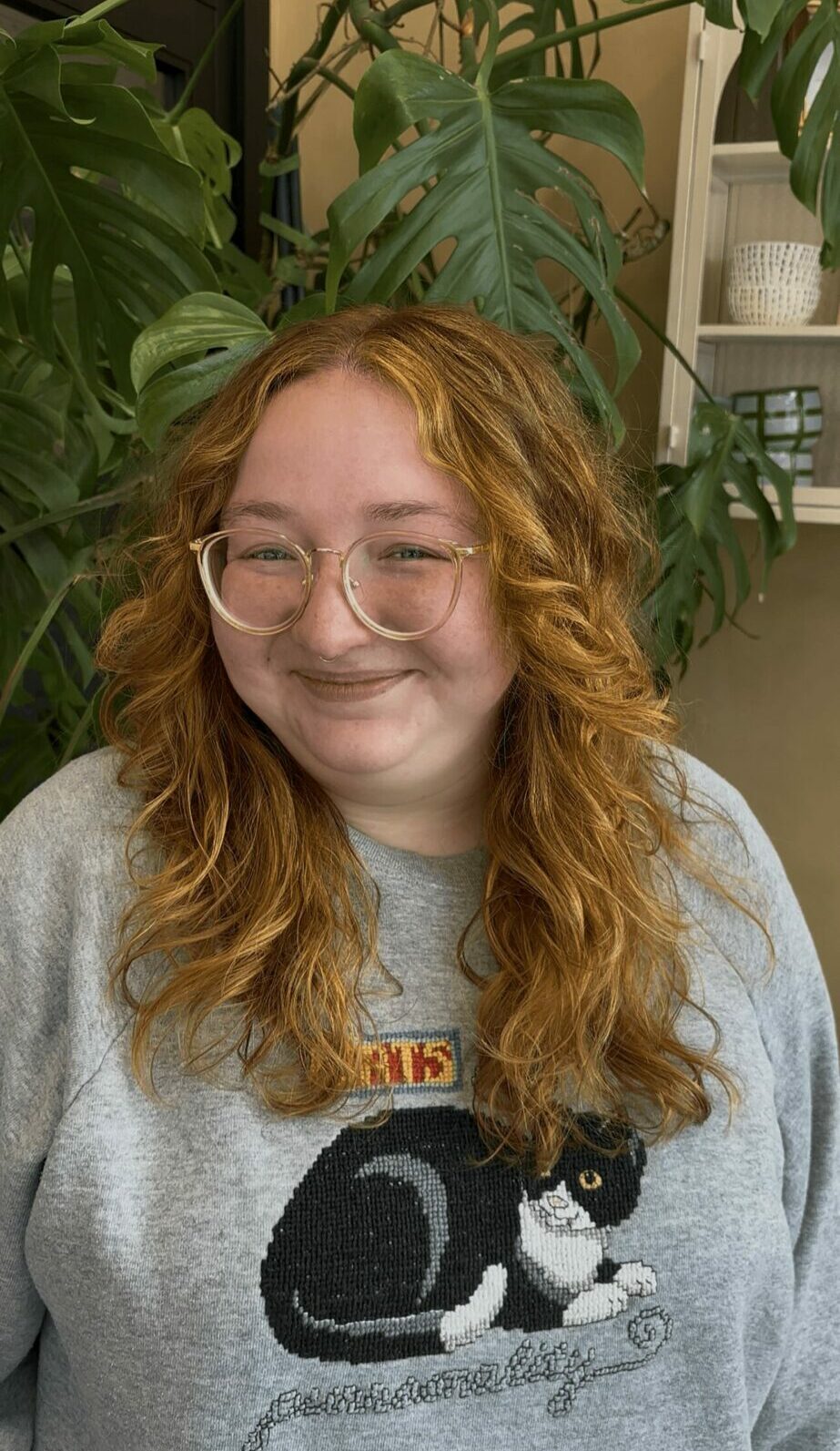 Young woman with curly red hair and glasses smiling, surrounded by large green plants indoors.