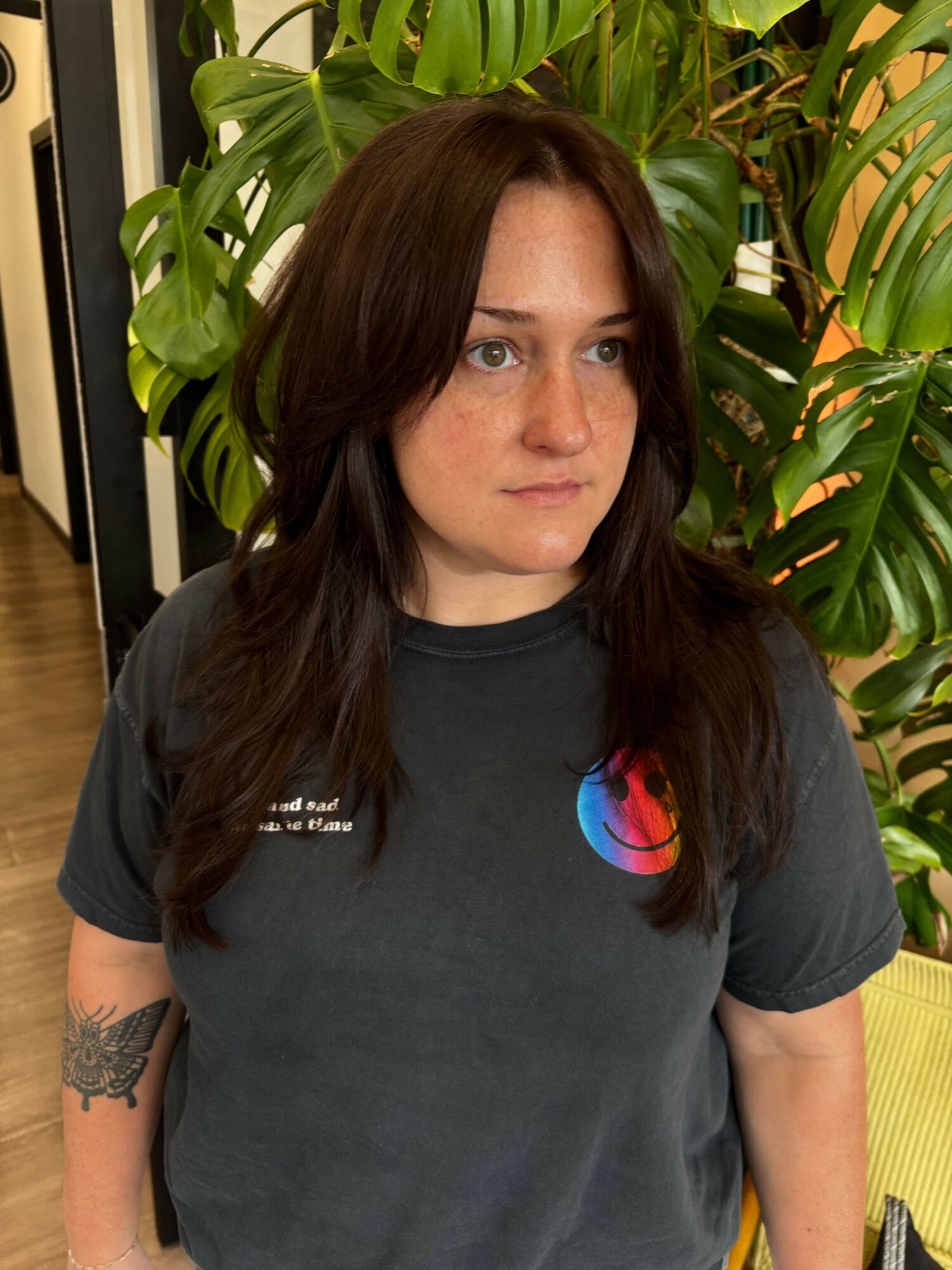 Woman with long dark hair lying among green tropical plants, wearing a black t-shirt with a colorful logo, looking at the camera.
