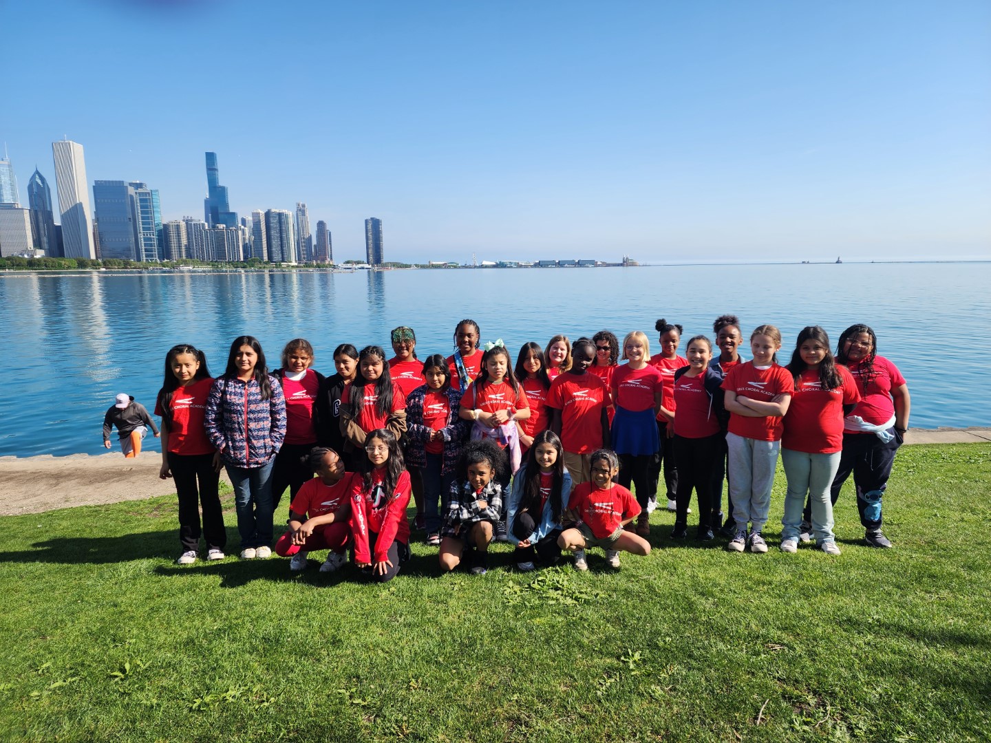 Group of children and adults standing on grass near water with city skyline in background.