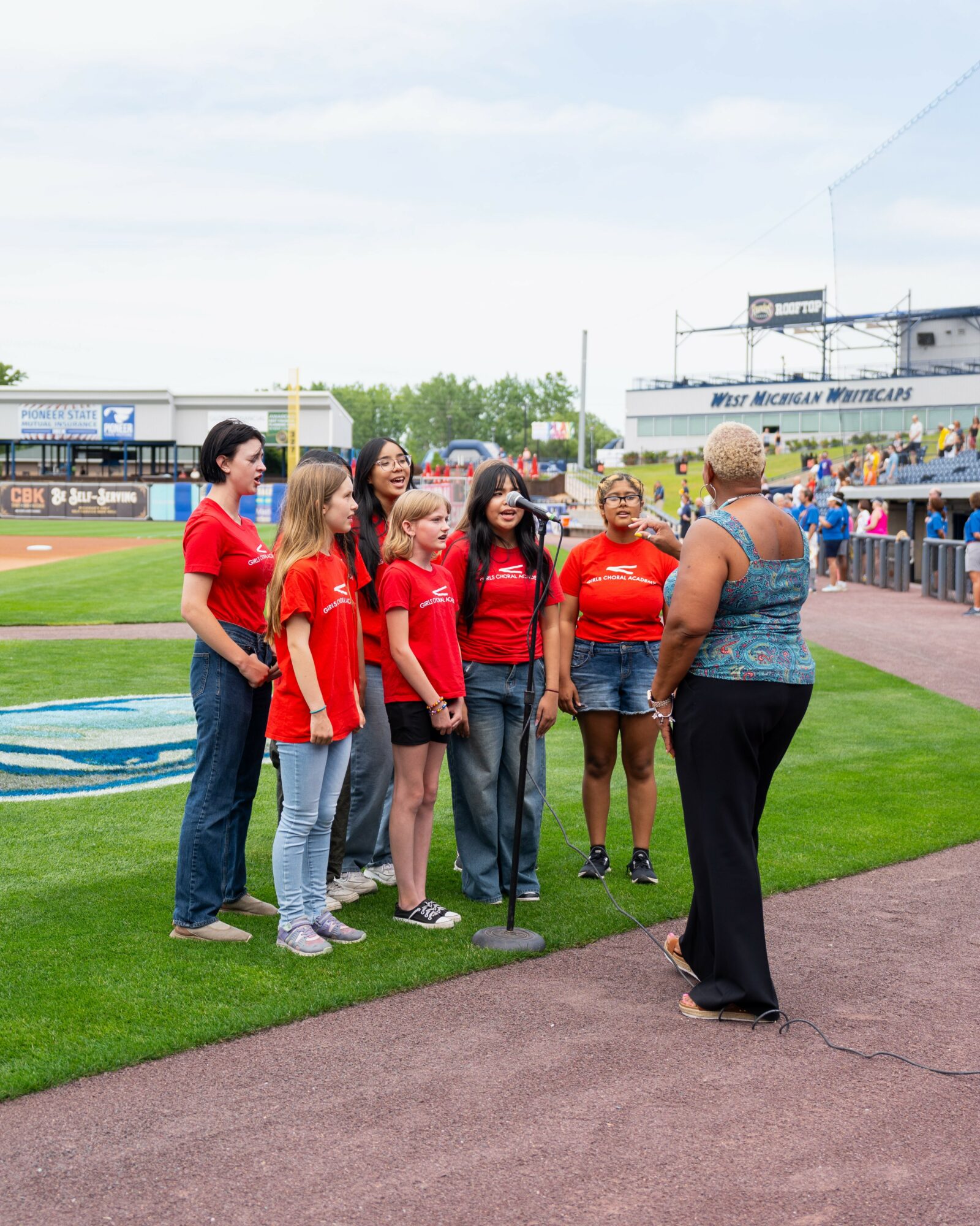 Group of six women in red shirts standing on a sports field, talking to a woman in a blue top, with stadium and sky in background.