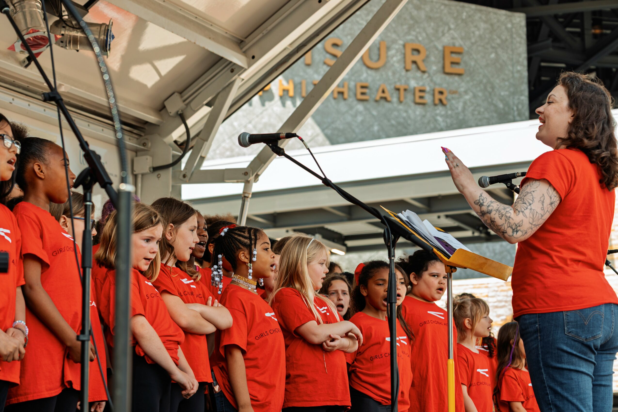 Group of children in red shirts listening to a woman speaking on stage outdoors.