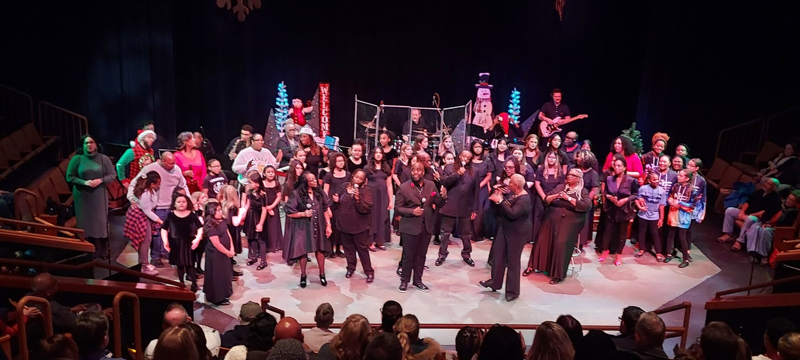 Group of people on stage performing or celebrating, with festive decorations and audience watching, in a theater setting.