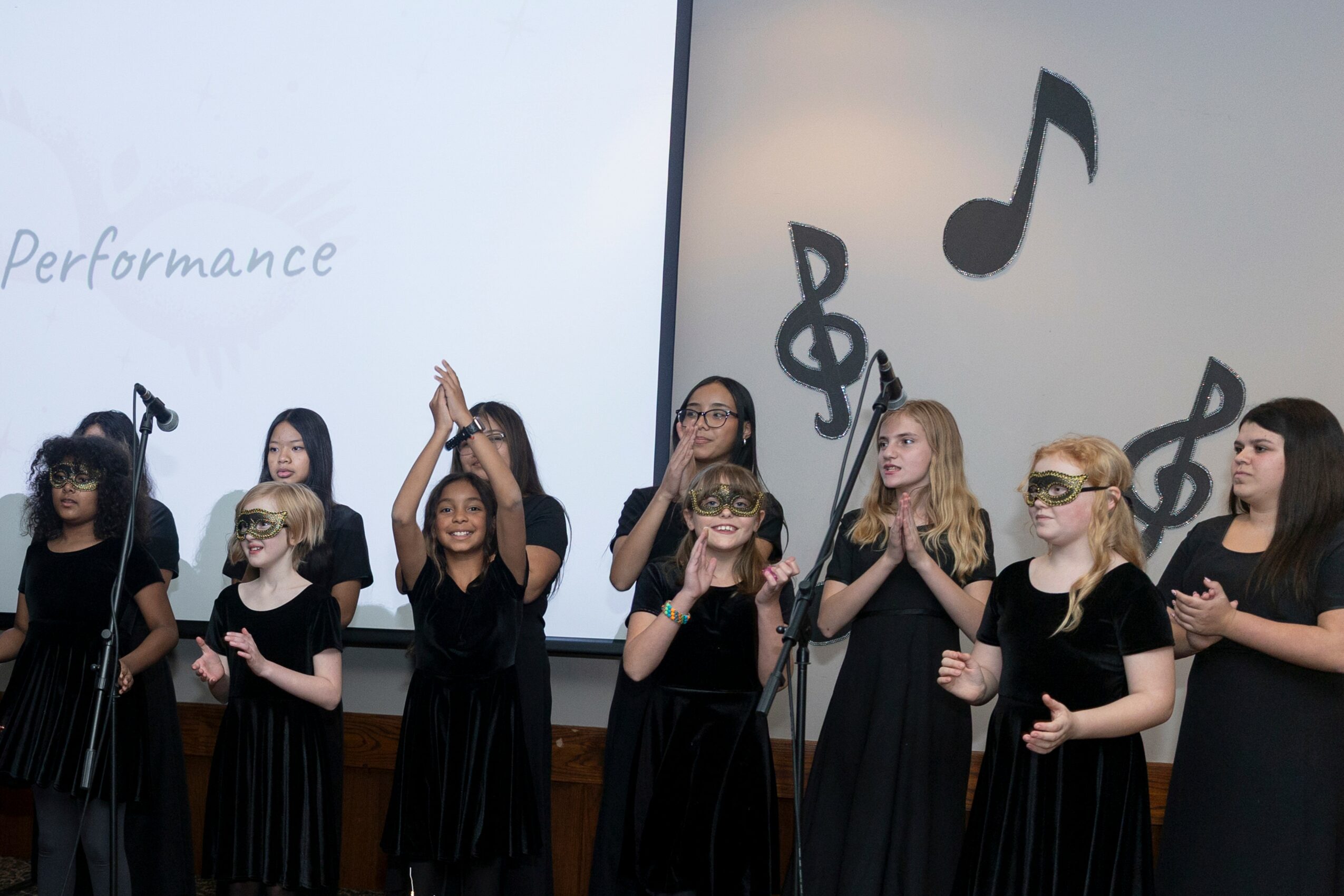 Group of children in black dresses performing on stage with musical note decorations, some wearing glasses, in front of a screen.