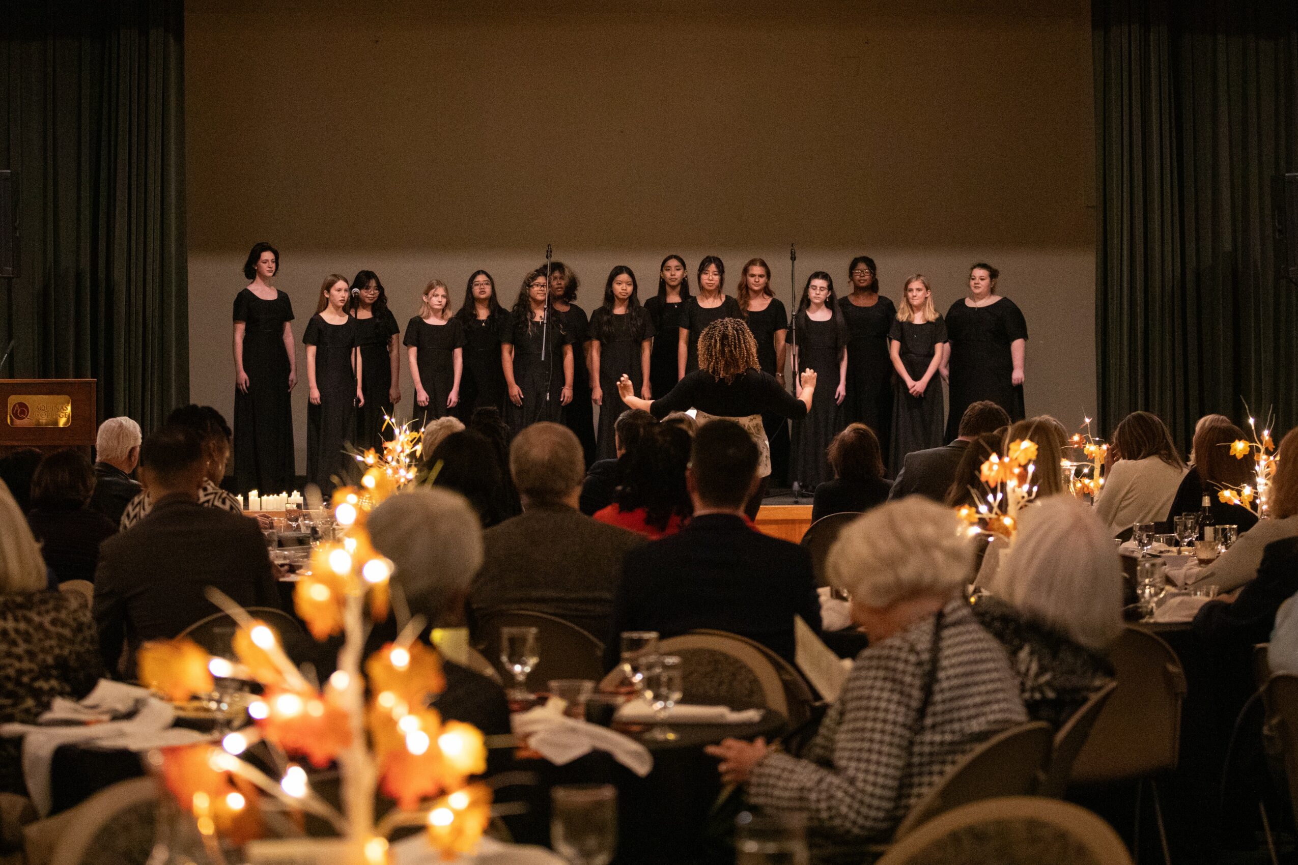 Chorus of women singing on stage in front of an audience at a formal event, with tables and decorations visible.