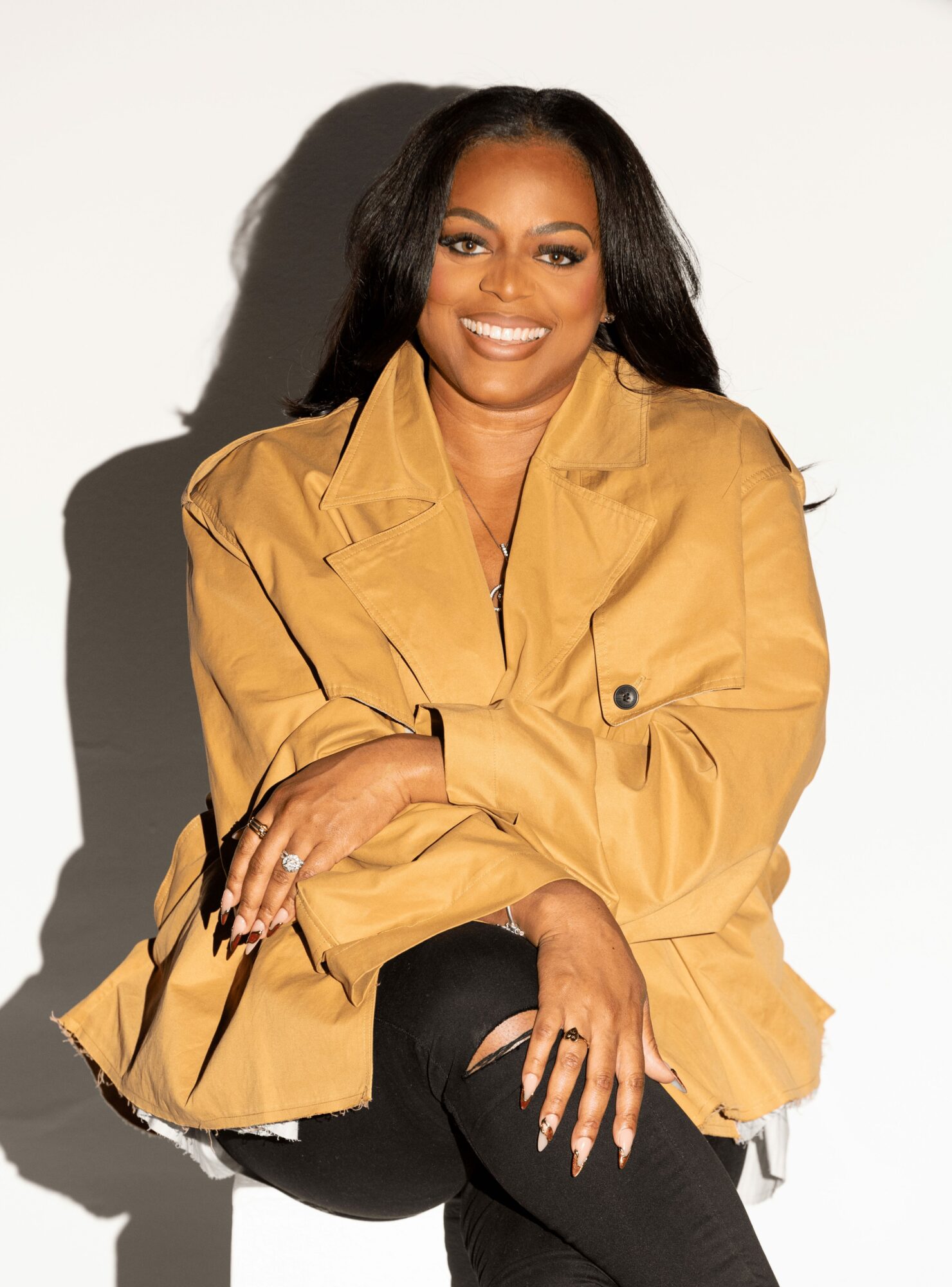Woman with long dark hair smiling, wearing a tan jacket and black pants, sitting against a white background.