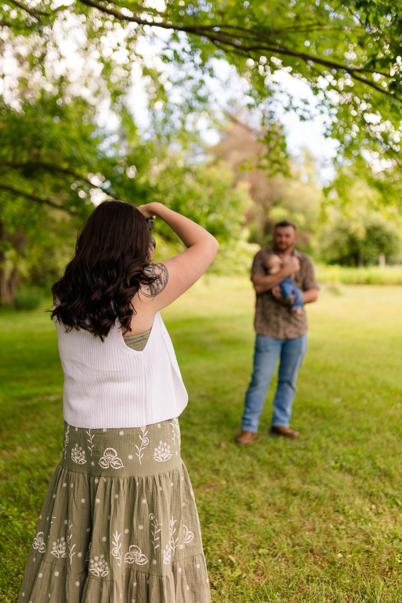 Woman with dark hair in a white top and patterned skirt stands outdoors, facing a man in a park with trees and grass