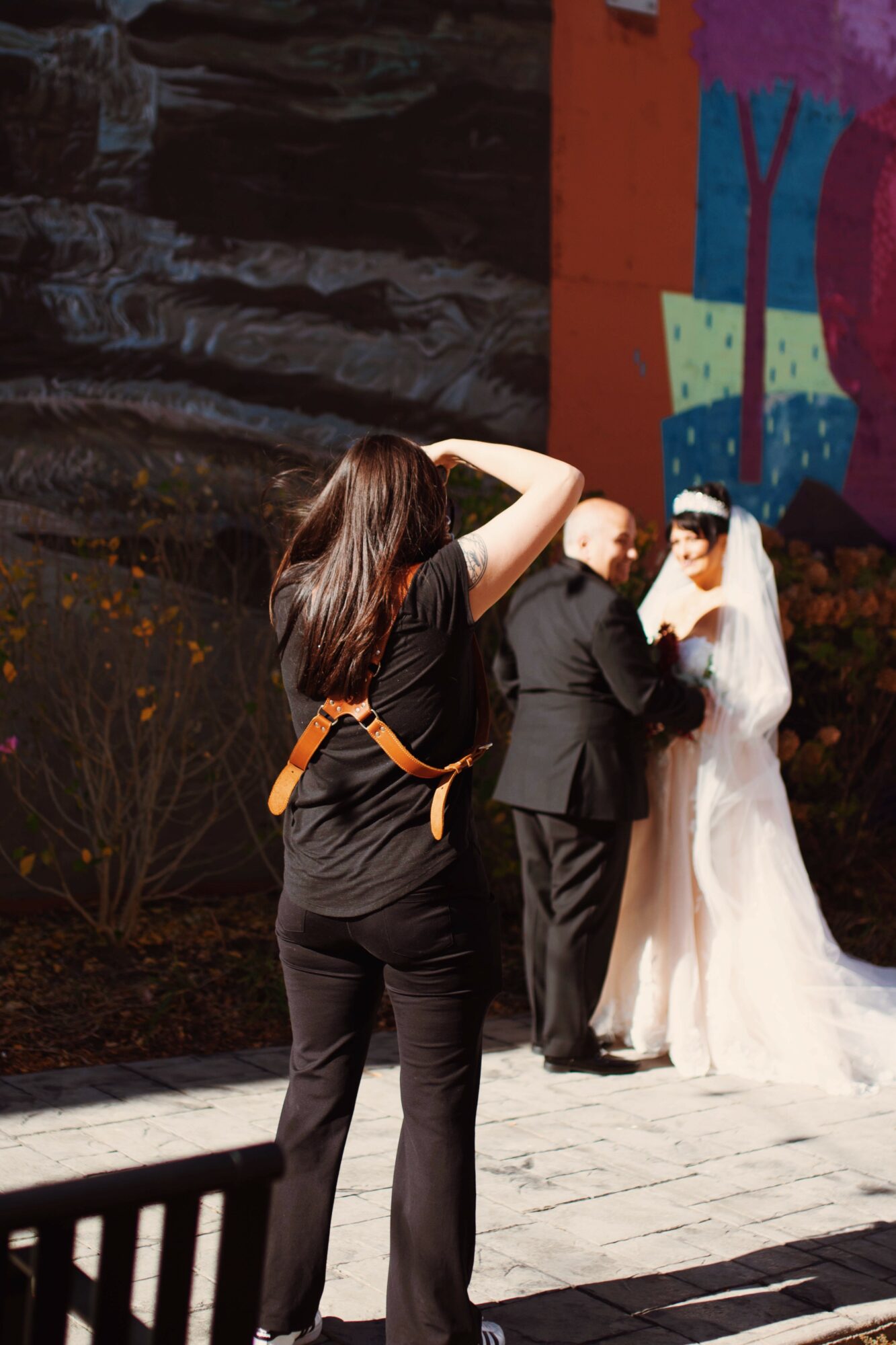 Person with long hair taking photo of a bride and groom outdoors, colorful backdrop behind them.