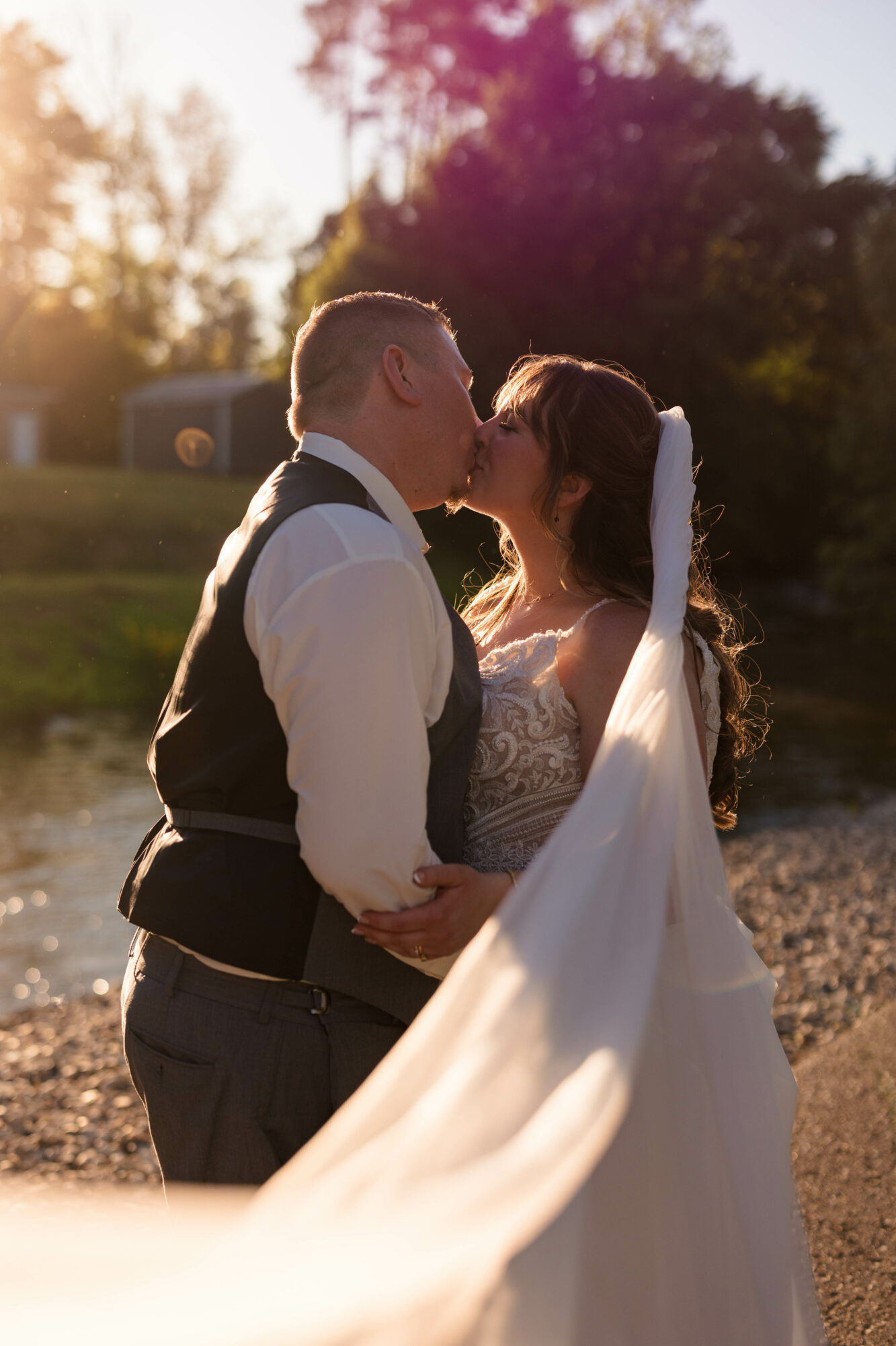 Couple kissing outdoors during sunset, with trees and sunlight in the background, bride in white dress and veil, groom in vest and shirt.