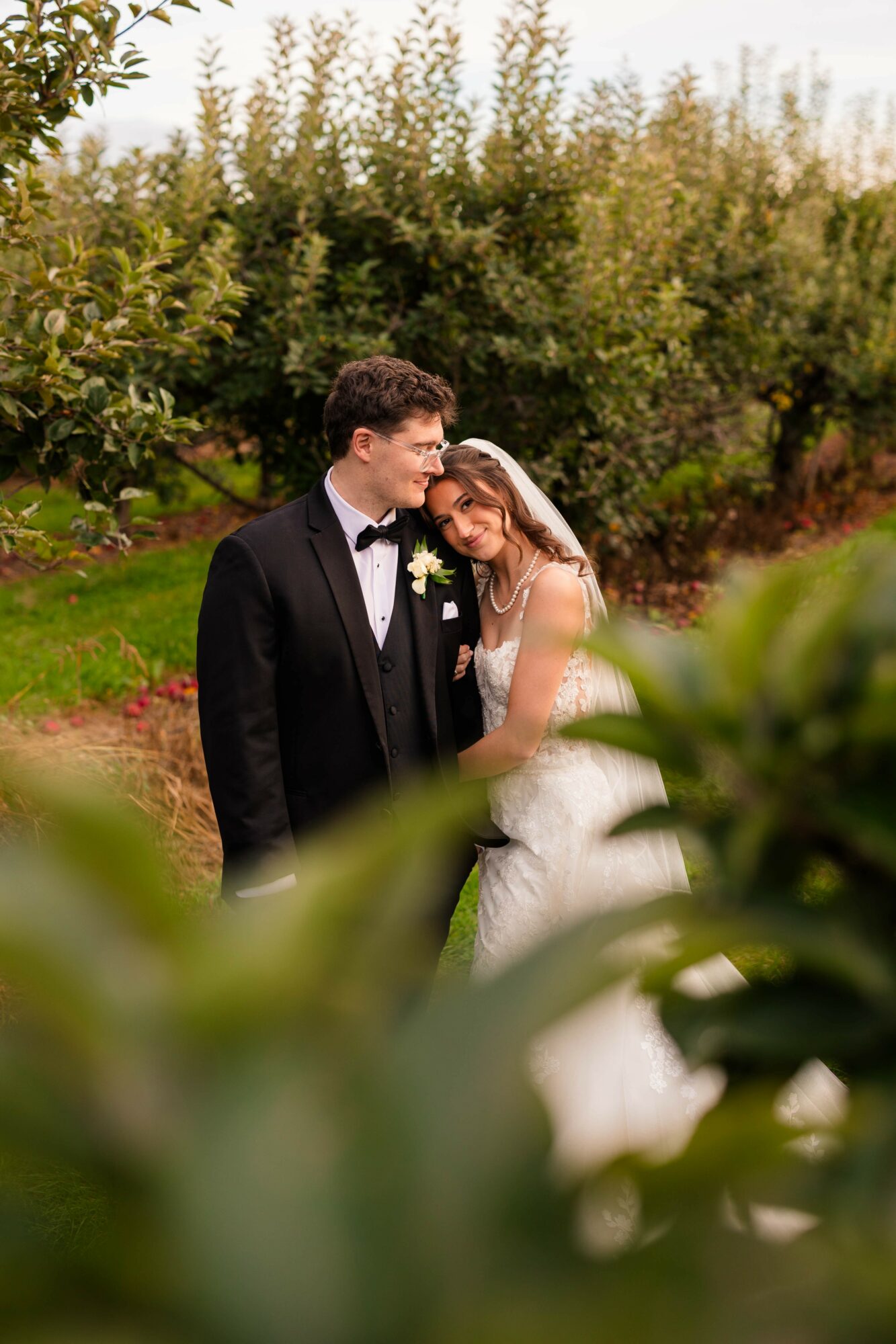 A bride and groom stand close together outdoors, surrounded by greenery and trees, during daytime. The bride leans her head on the groom's shoulder.
