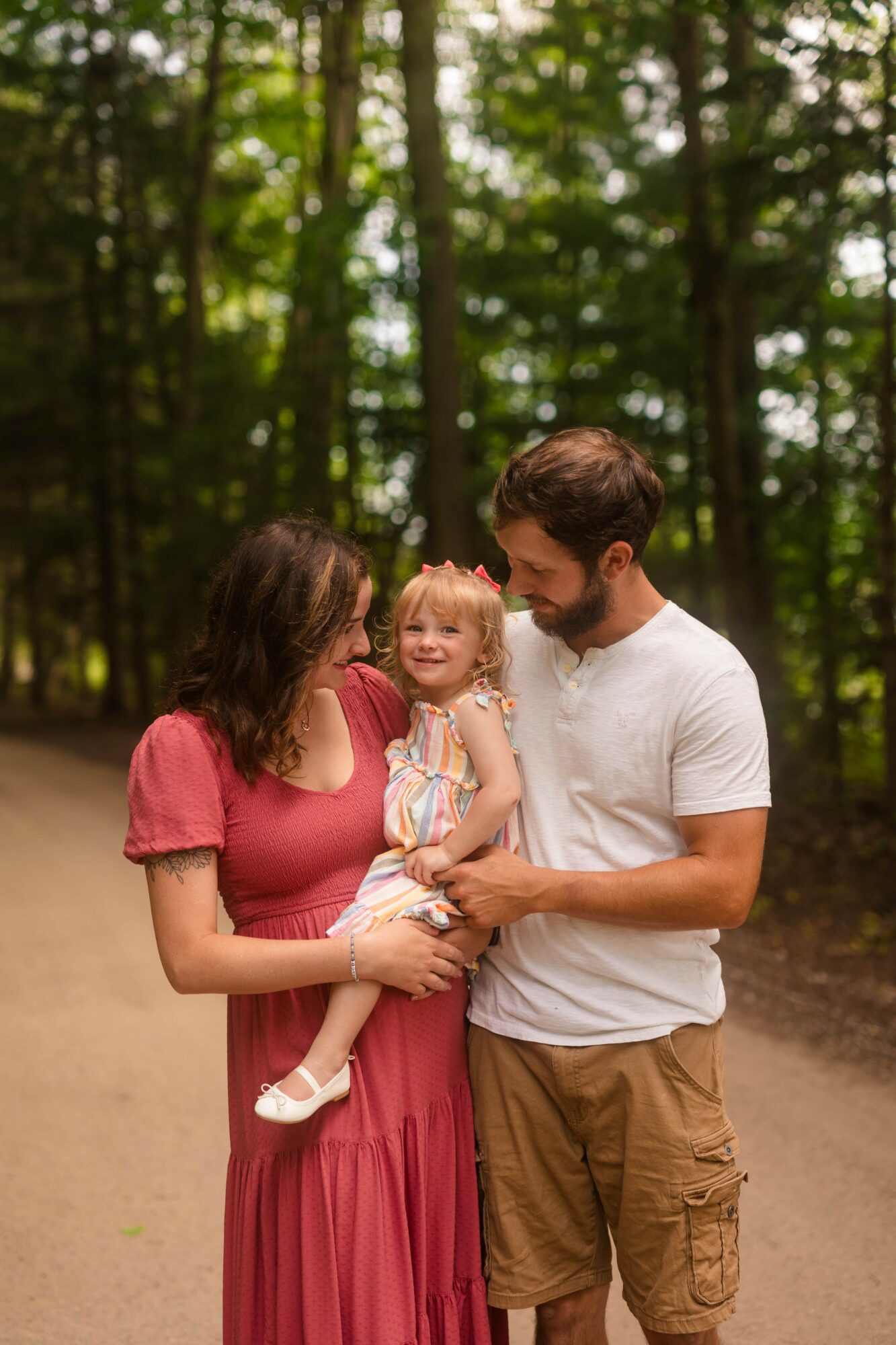 Family of three standing on a forest path, smiling and looking at each other, surrounded by trees.