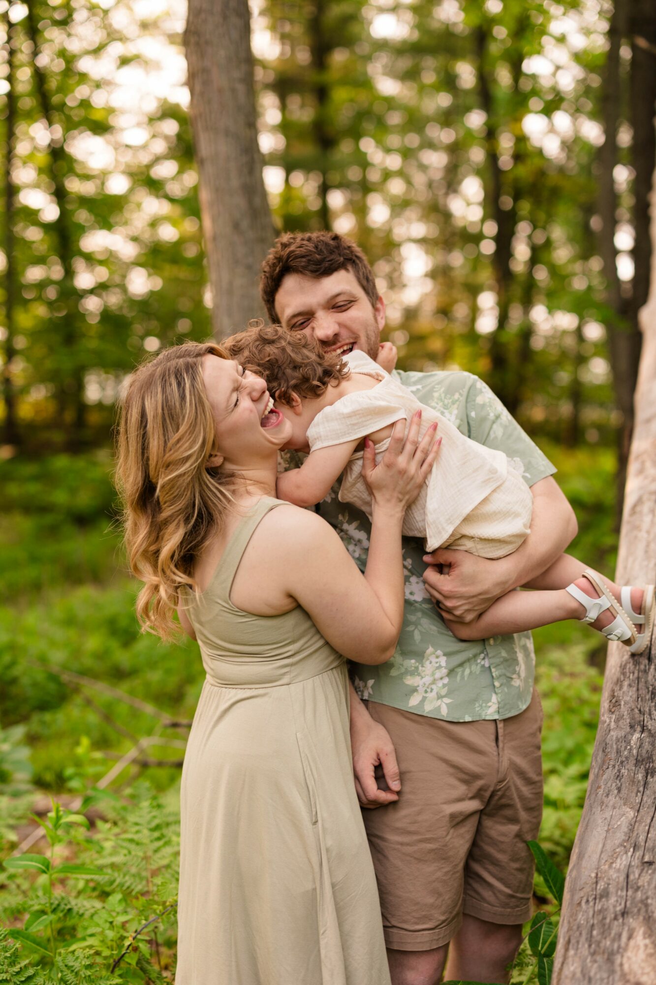 A smiling couple holding a baby outdoors in a forested area with trees and greenery.