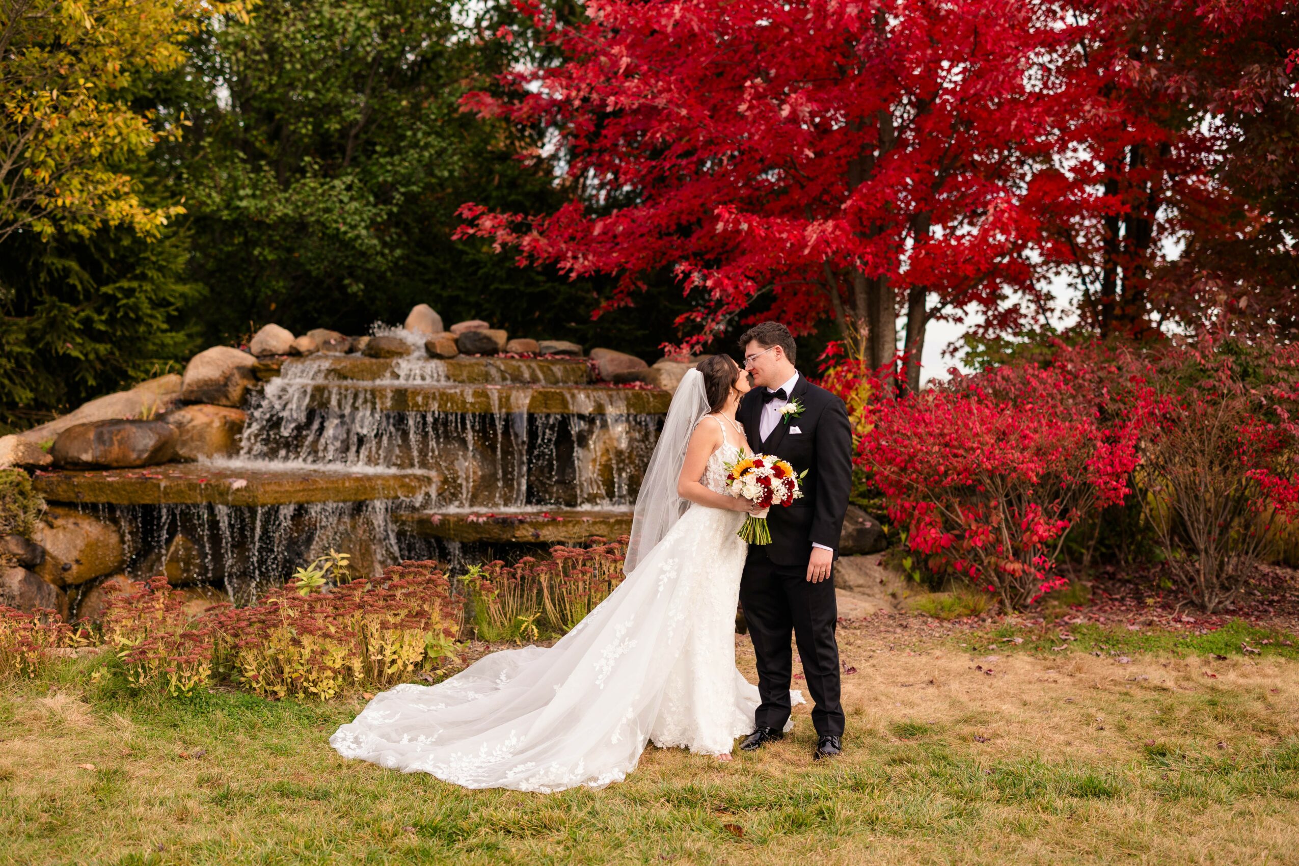 Bride and groom stand close together outdoors with colorful autumn trees and a waterfall in the background.