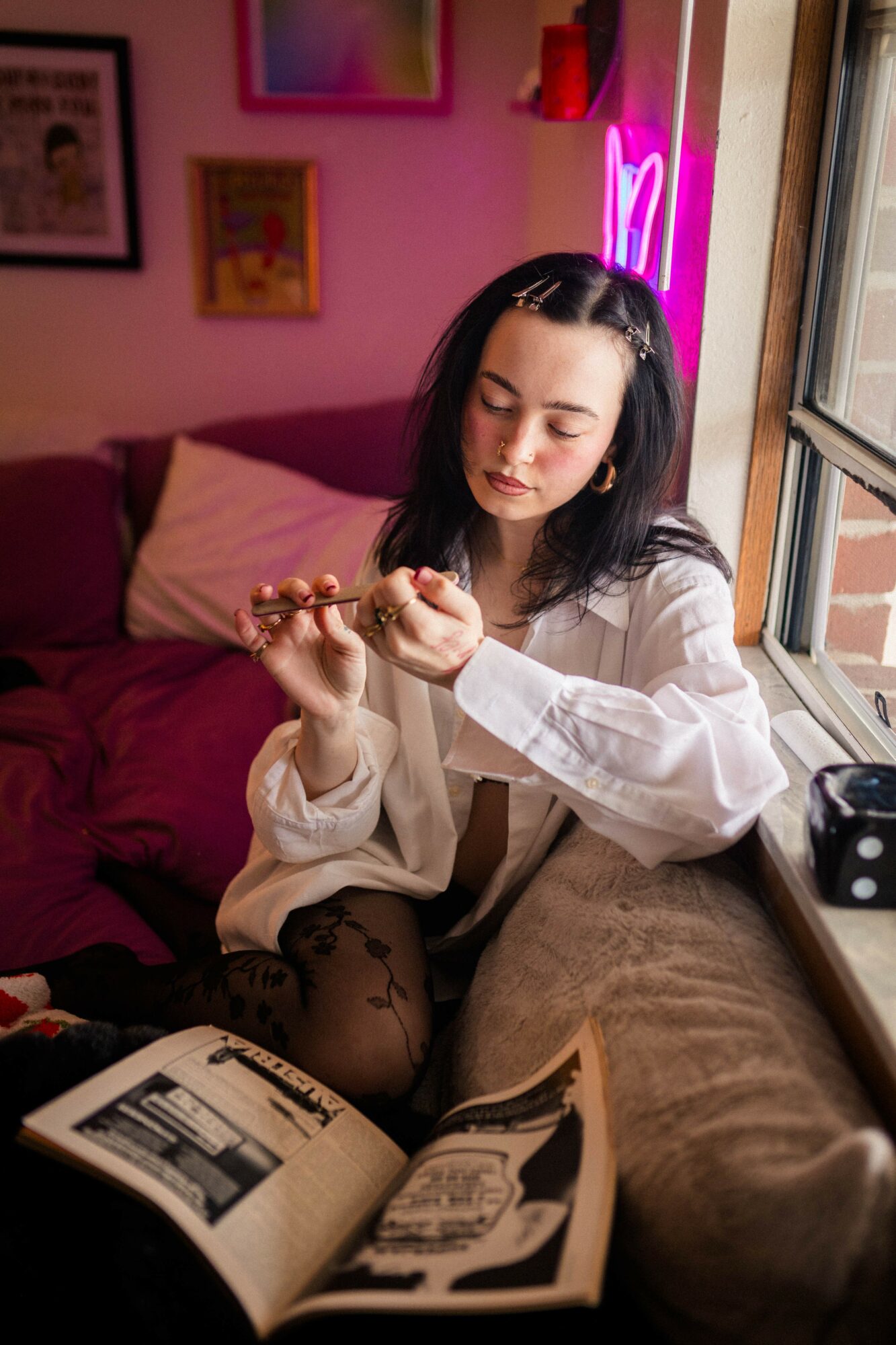 Young woman with dark hair sitting by a window, reading a book, with pink neon lights in the background.