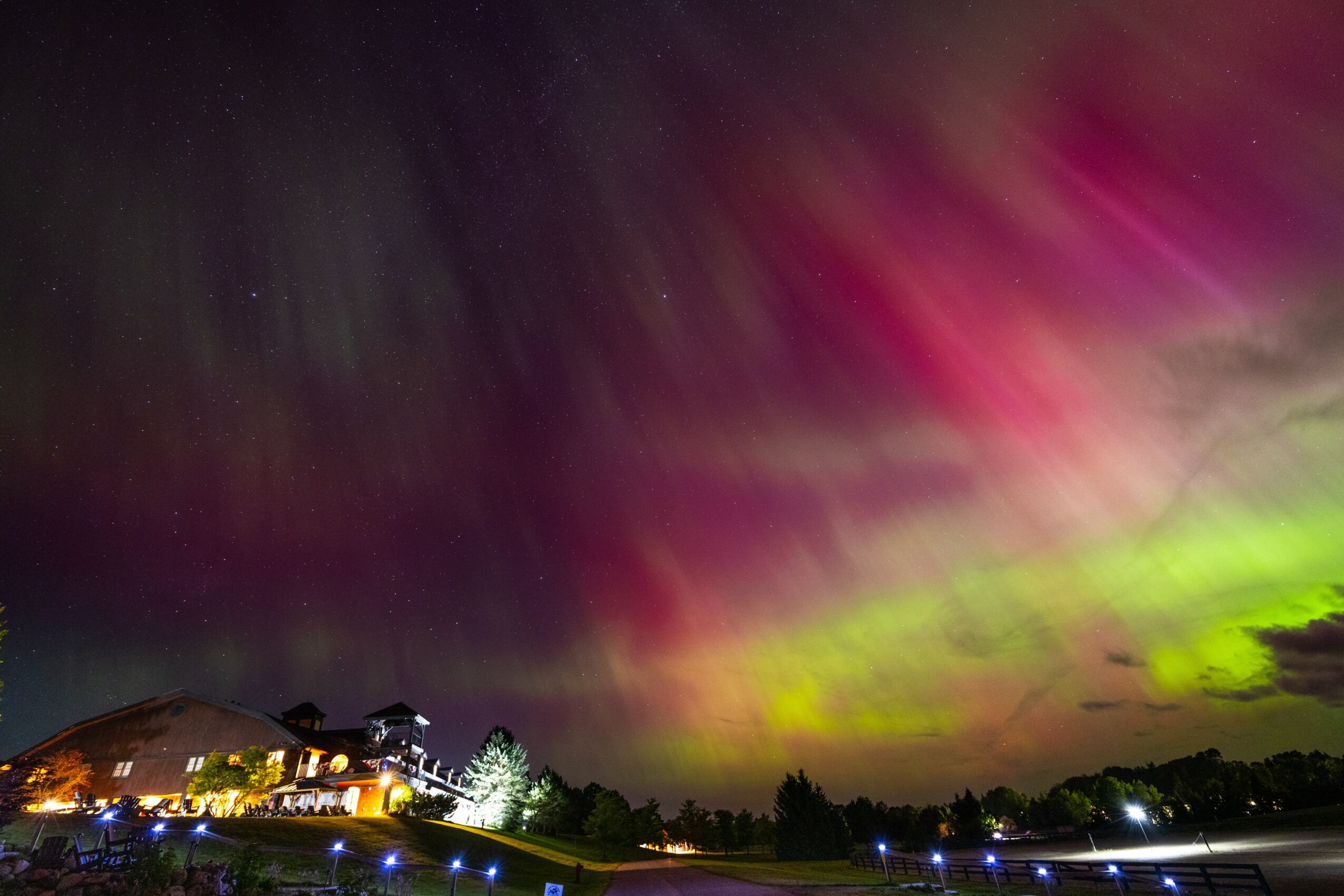 Night sky with colorful aurora borealis, green, pink, and purple hues, above a landscape with illuminated buildings and trees.