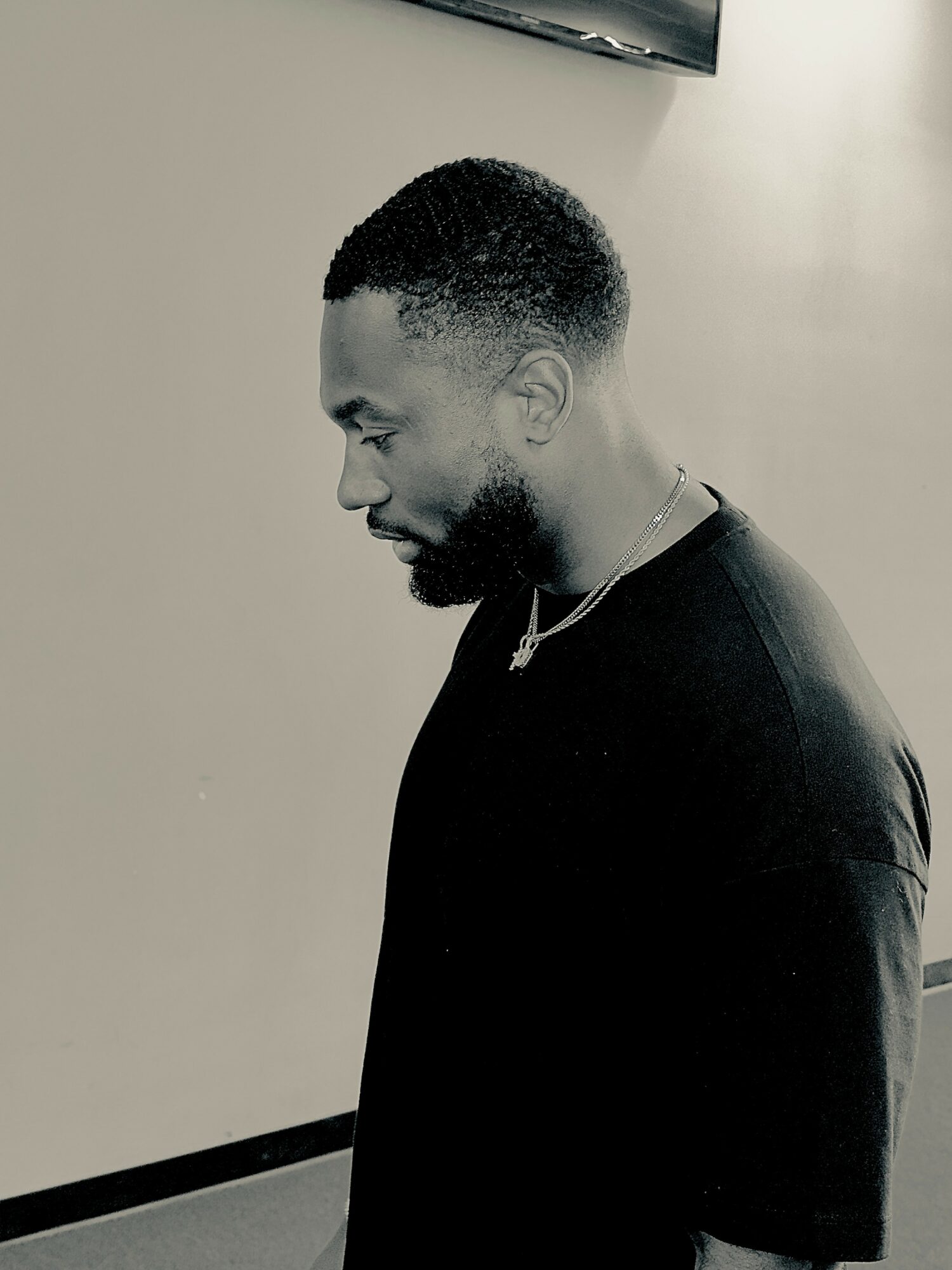 Side profile of a man with short curly hair and a beard, wearing a dark shirt, looking downward.