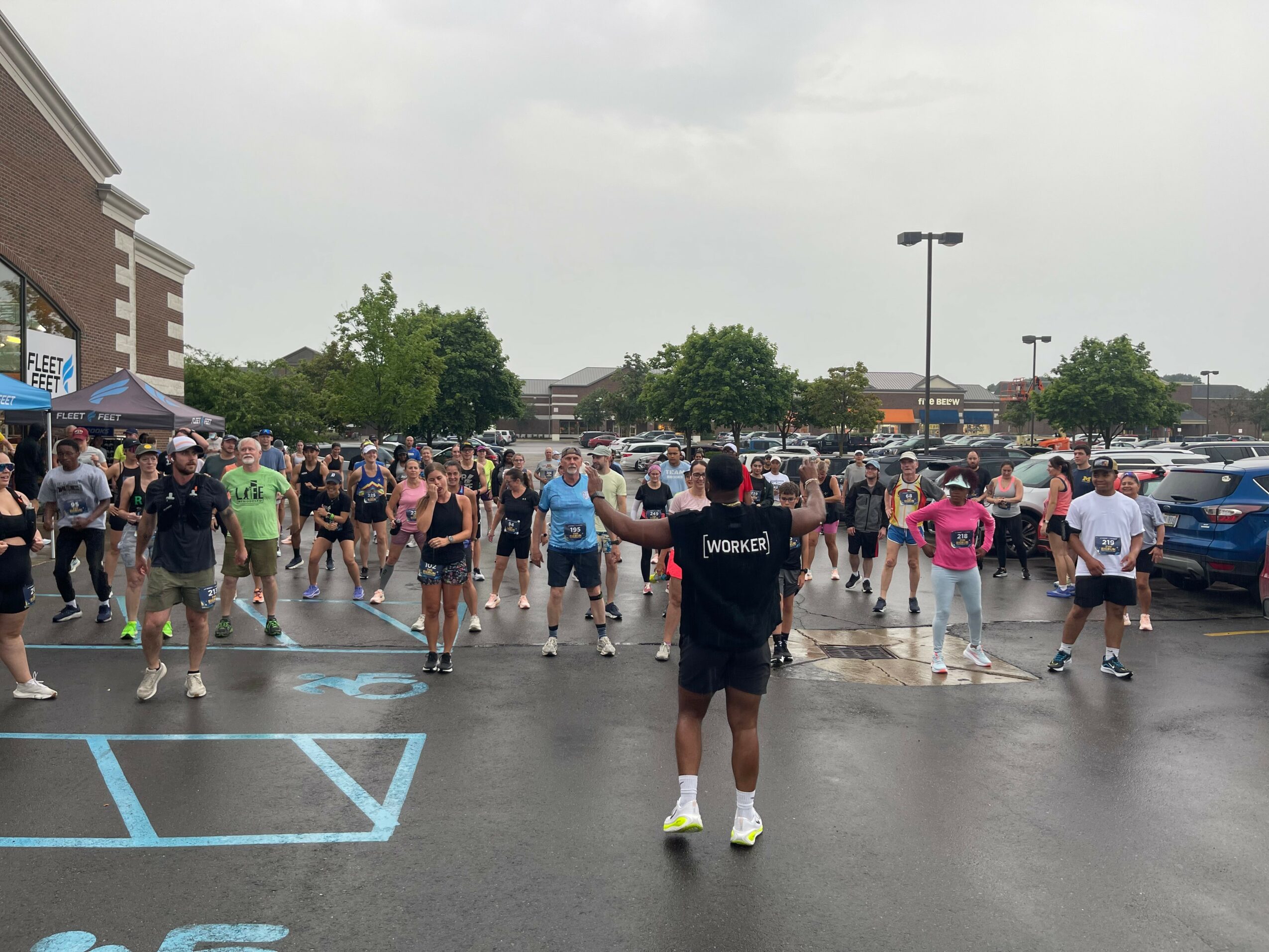 Group of people gathered in a parking lot, some walking, some standing, with cars and buildings in the background.
