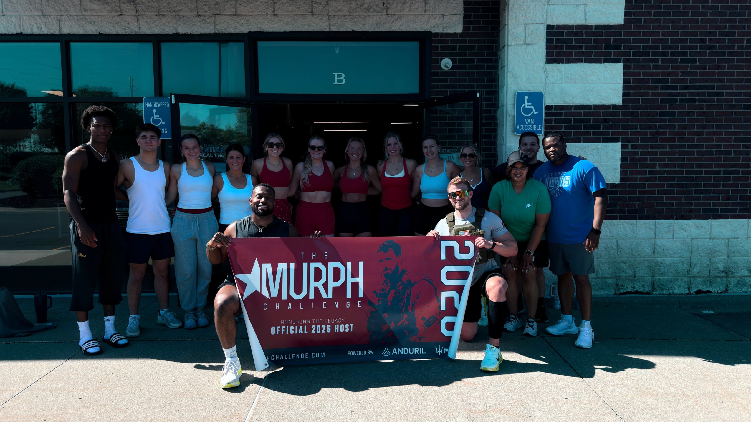 Group of people holding a banner outside a building, some wearing athletic clothing, with a blue sign and brick wall behind.