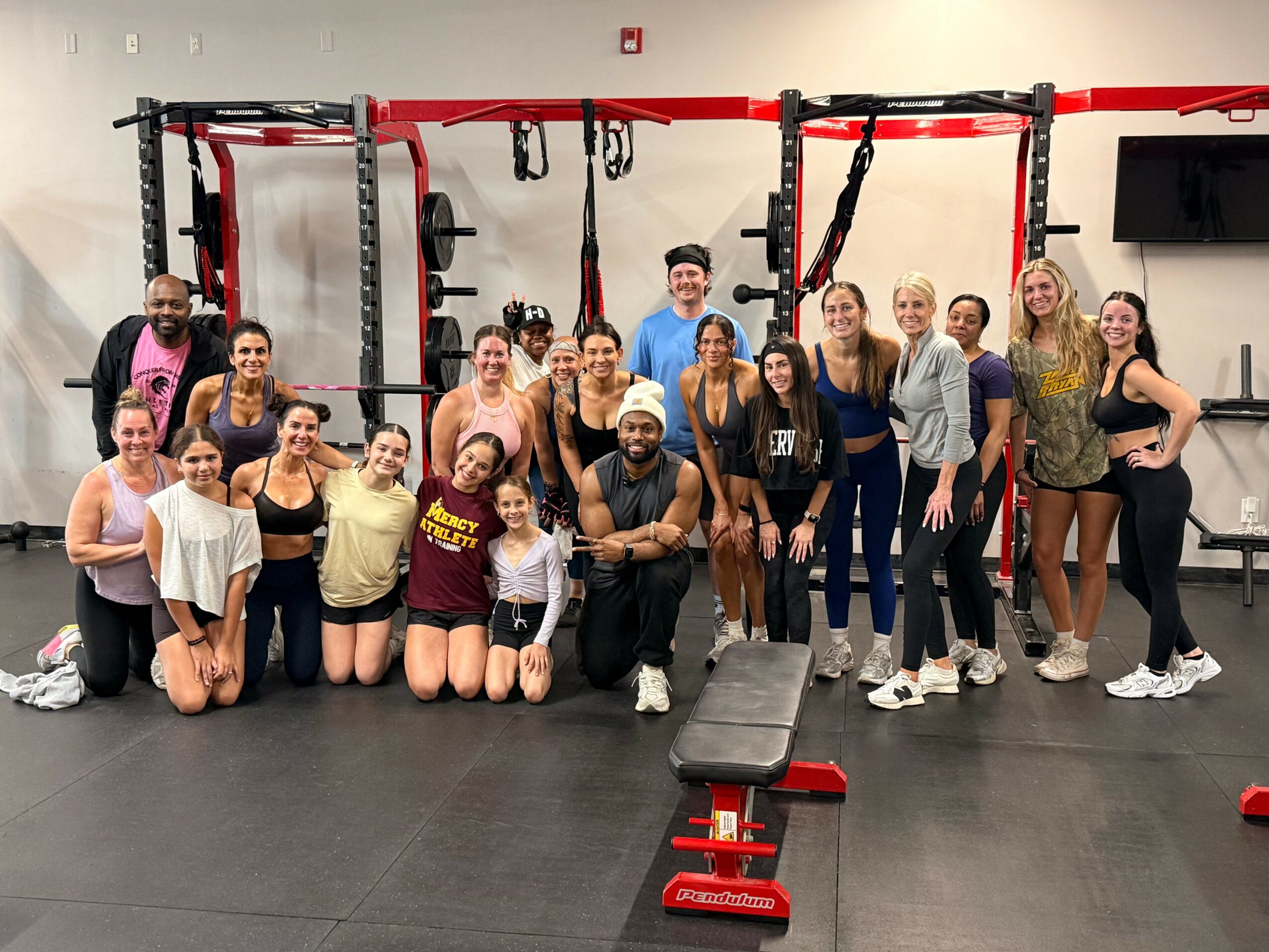 Group of people in a gym, some kneeling and some standing, with gym equipment in the background.