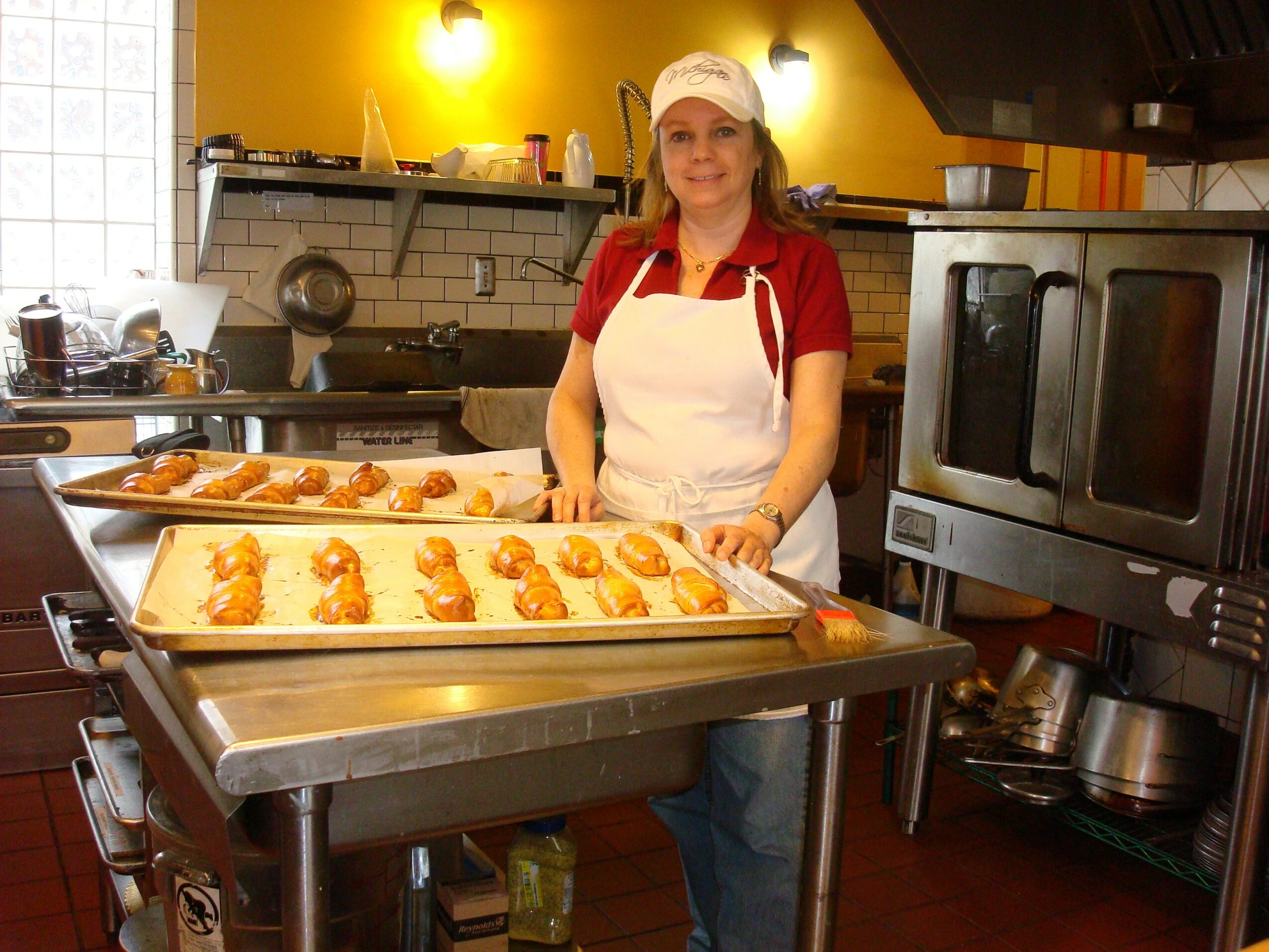 Woman in a kitchen wearing a white apron and cap, standing behind trays of baked goods, with kitchen appliances around.