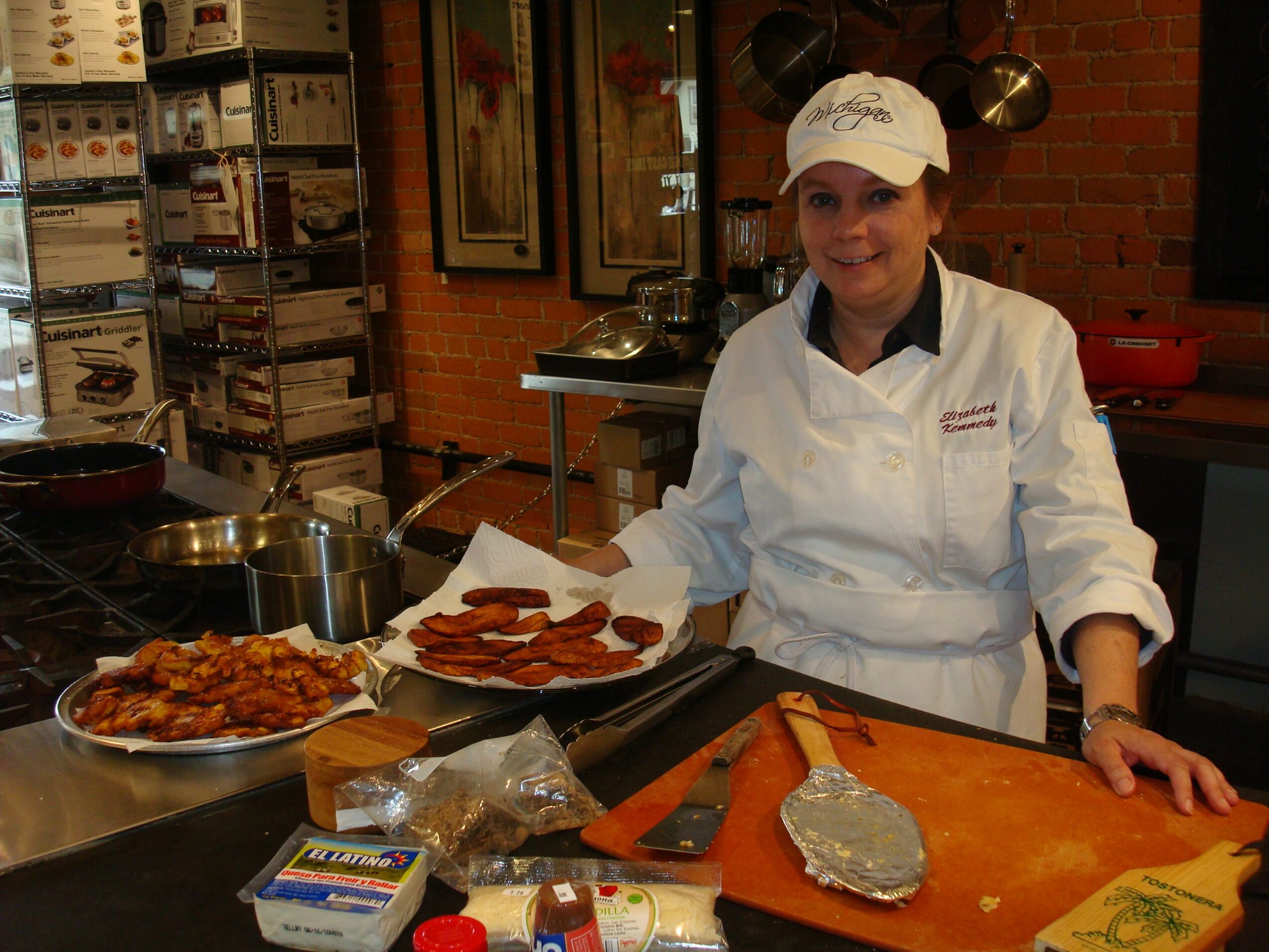 Chef in white uniform and cap standing behind a kitchen counter with prepared food, cutting board, and cooking utensils.