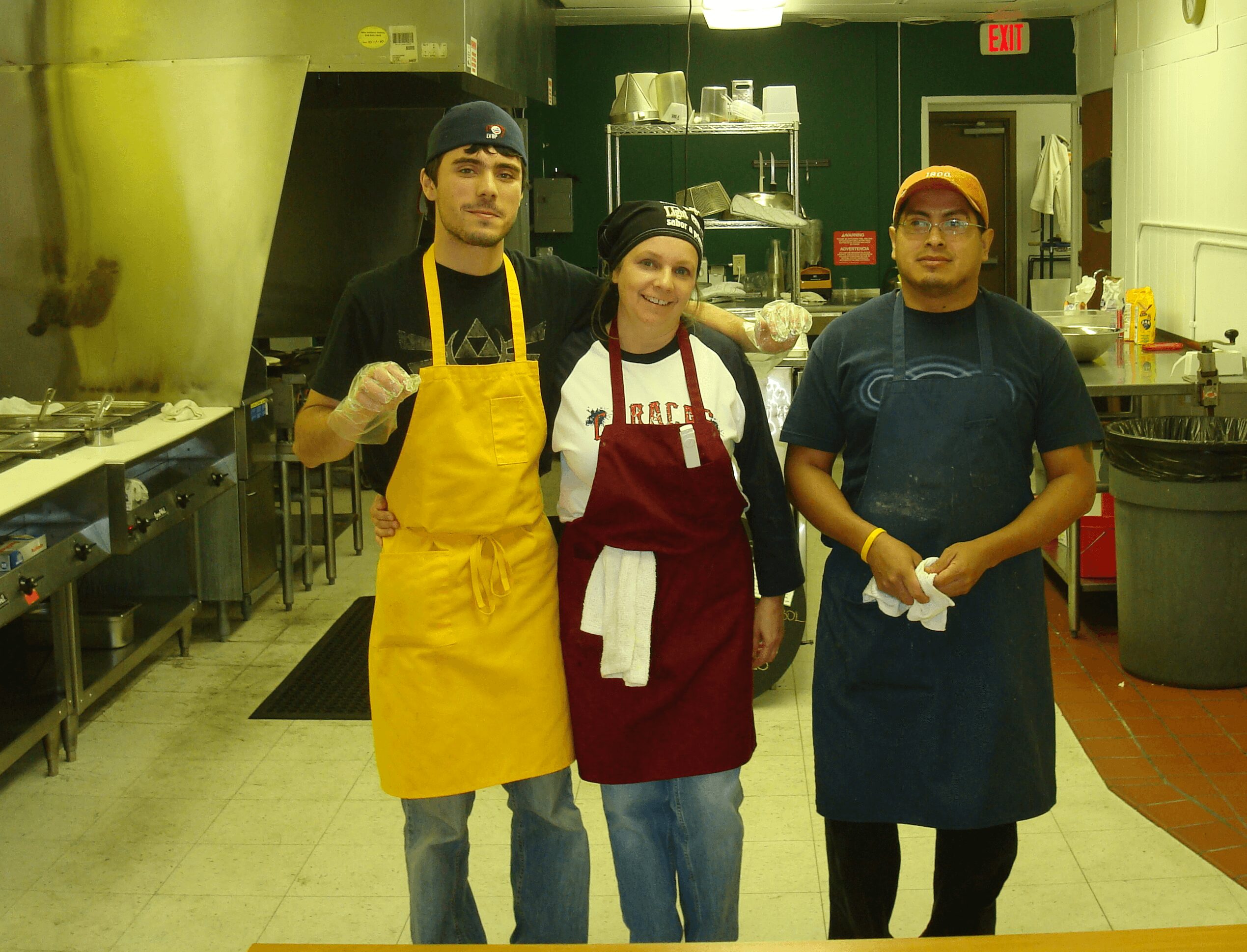 Three people in a kitchen, wearing aprons and gloves, standing side by side, smiling at the camera.