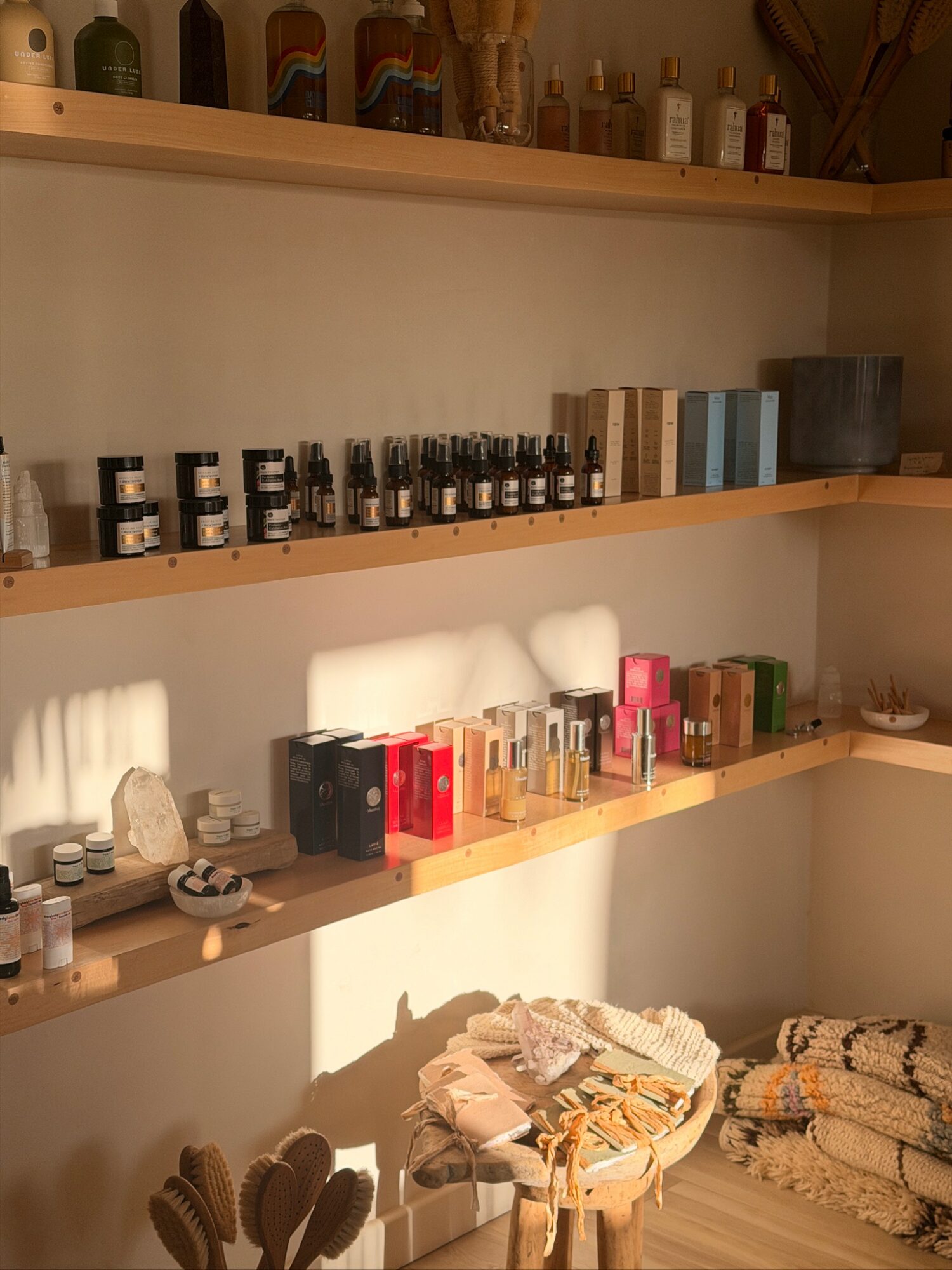 Shelves with bottles, boxes, and containers, with sunlight casting shadows on the wall, and a small table with folded towels.