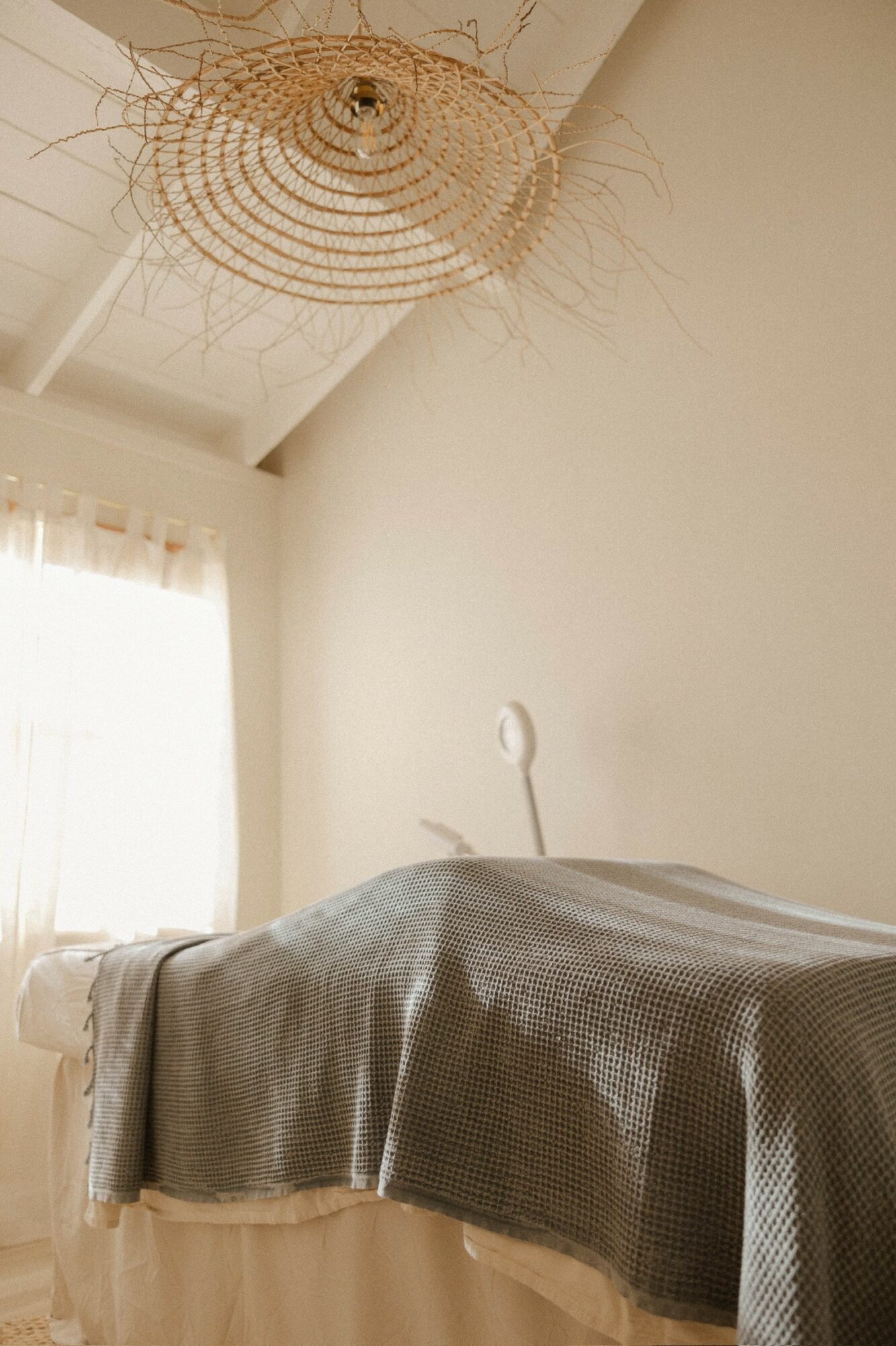 Bedroom with a bed covered in a textured gray blanket, a white wall, and a woven pendant light fixture.