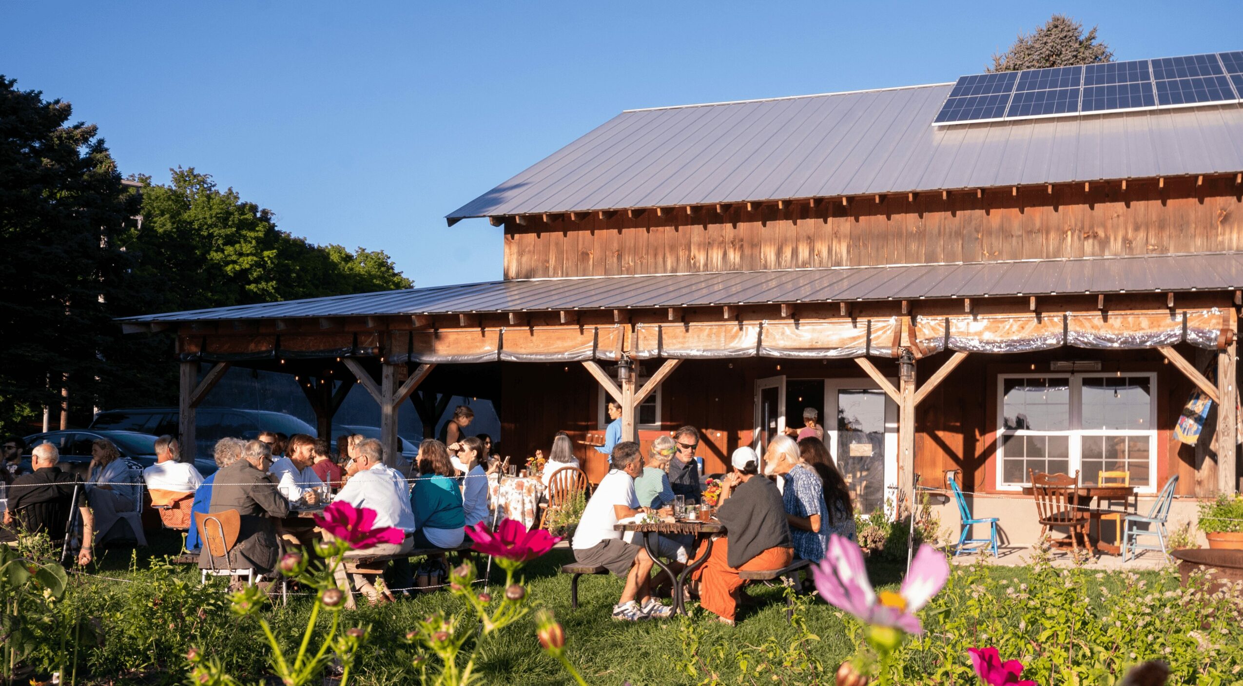 People sitting outdoors in front of a wooden building with solar panels on the roof, surrounded by flowers and greenery.