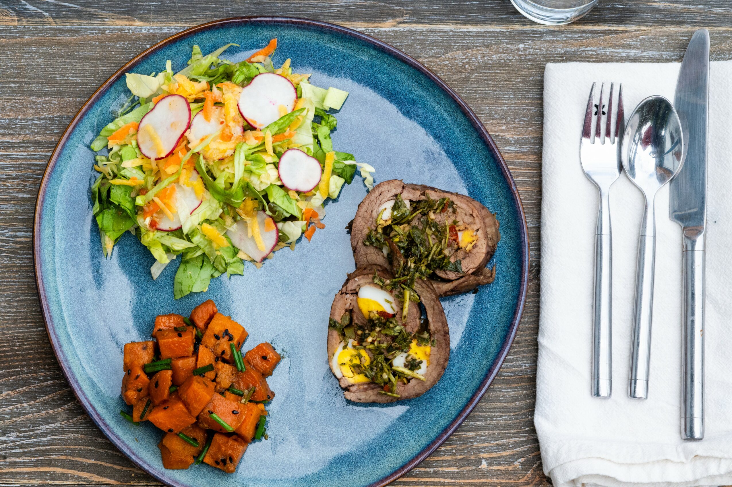 Plate with salad, two slices of meat with herbs, and diced sweet potatoes on a wooden table, with cutlery on a napkin.
