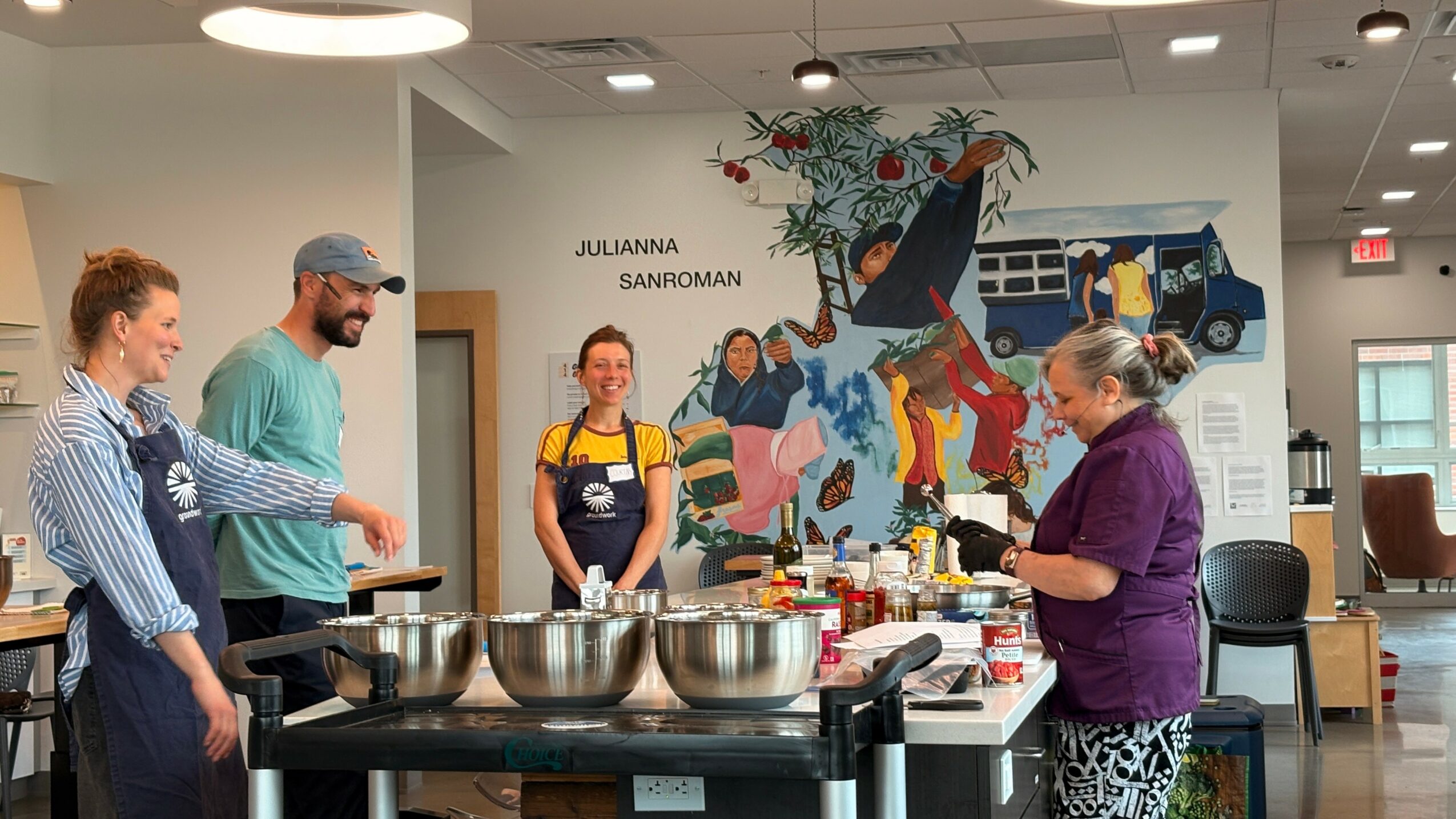 Four people in a room with a colorful mural, preparing food at a table with bowls and ingredients.