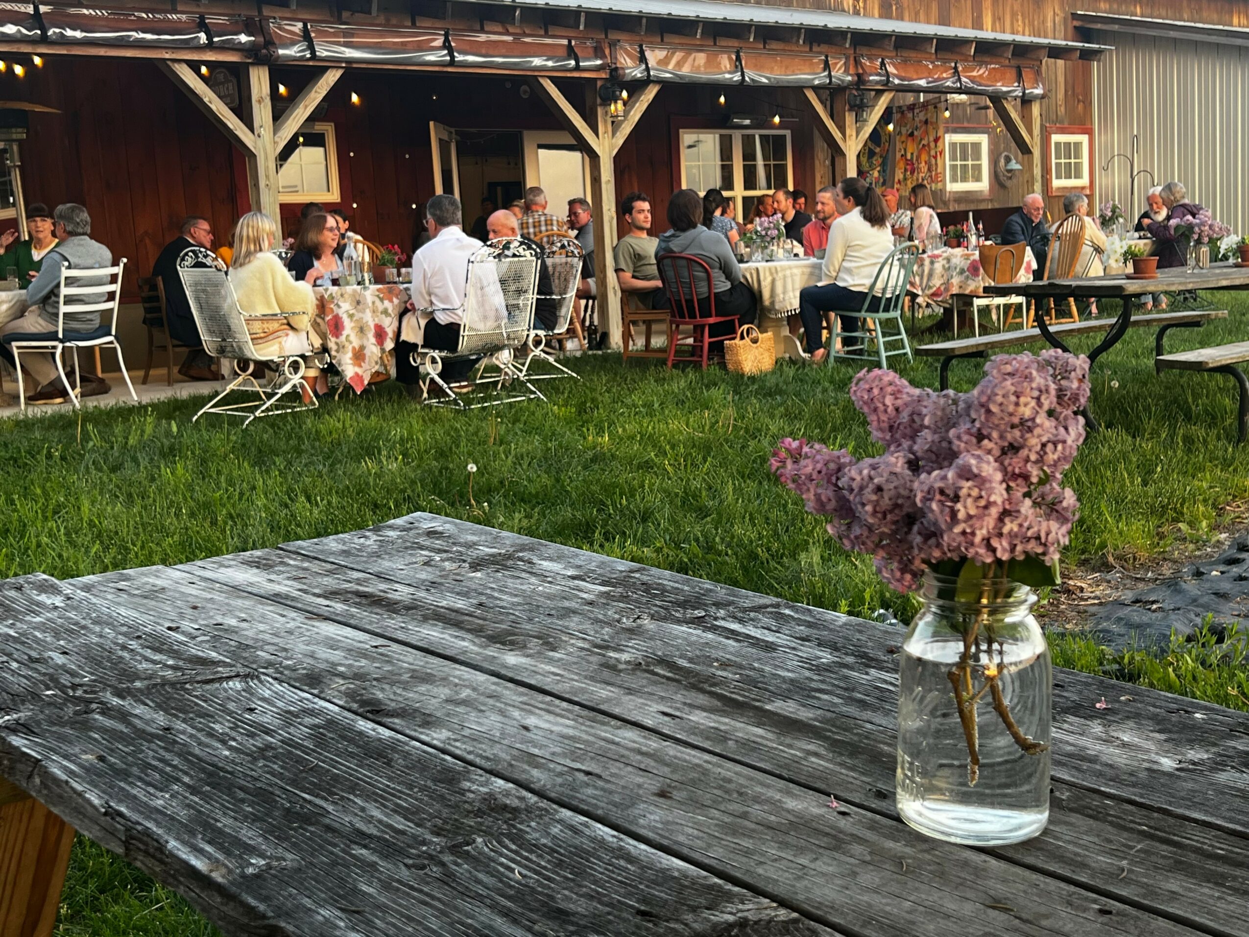 People dining outdoors at a gathering, with a rustic building in the background and a wooden table with a vase of pink flowers in the foreground.