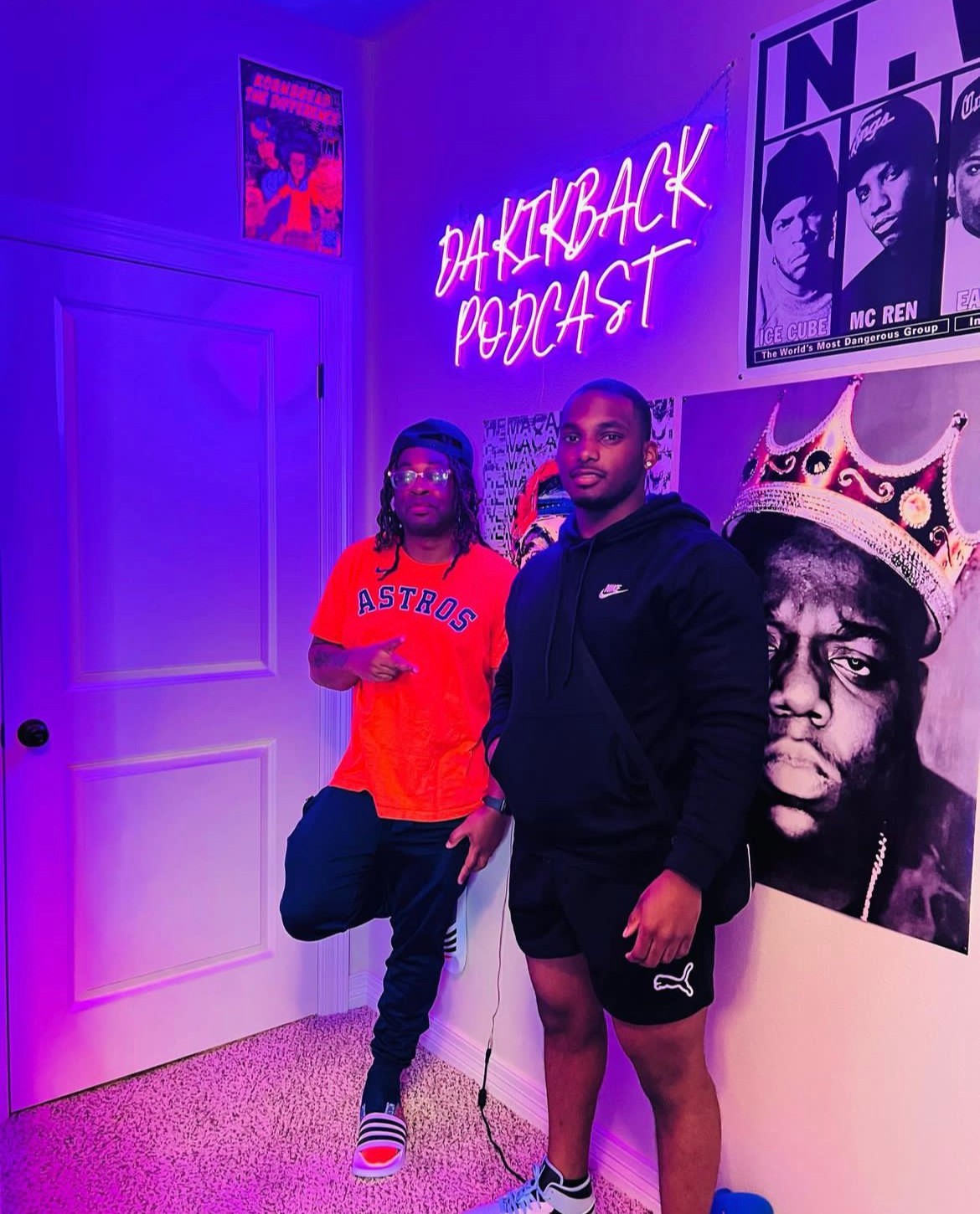 Two people standing in a room with neon signs and posters, one wearing a red shirt and the other in black, under purple lighting.