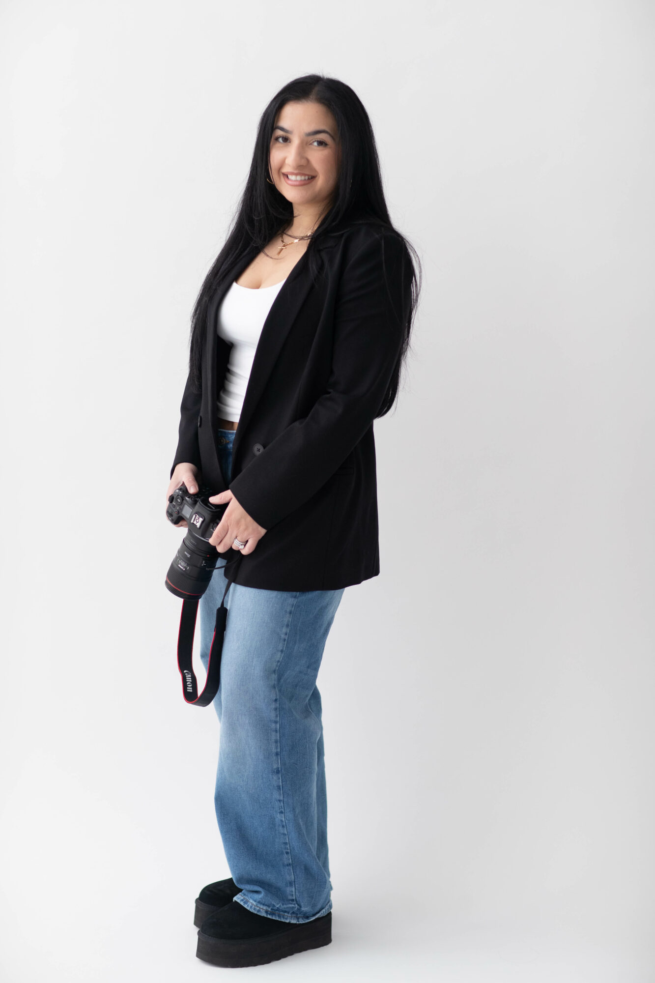 Young woman with long dark hair holding a camera, smiling, wearing a black blazer, white top, and blue jeans, standing against a white background.