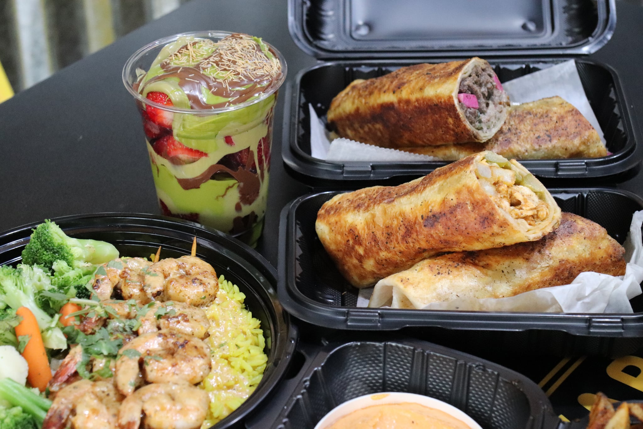 Two spring rolls in black containers, a salad with broccoli, carrots, and rice, and a cup with layered dessert with fruit and jelly.