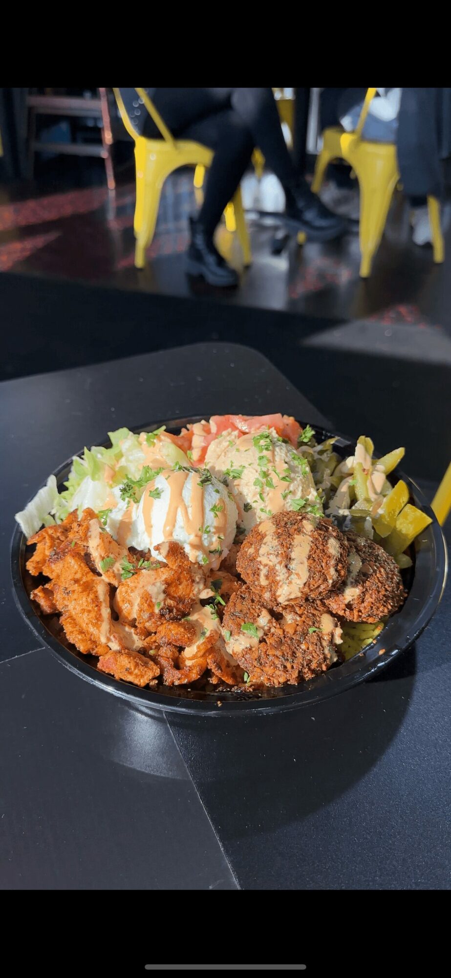 Bowl of fried chicken, lettuce, and toppings on a black table, with people sitting in yellow chairs in the background.