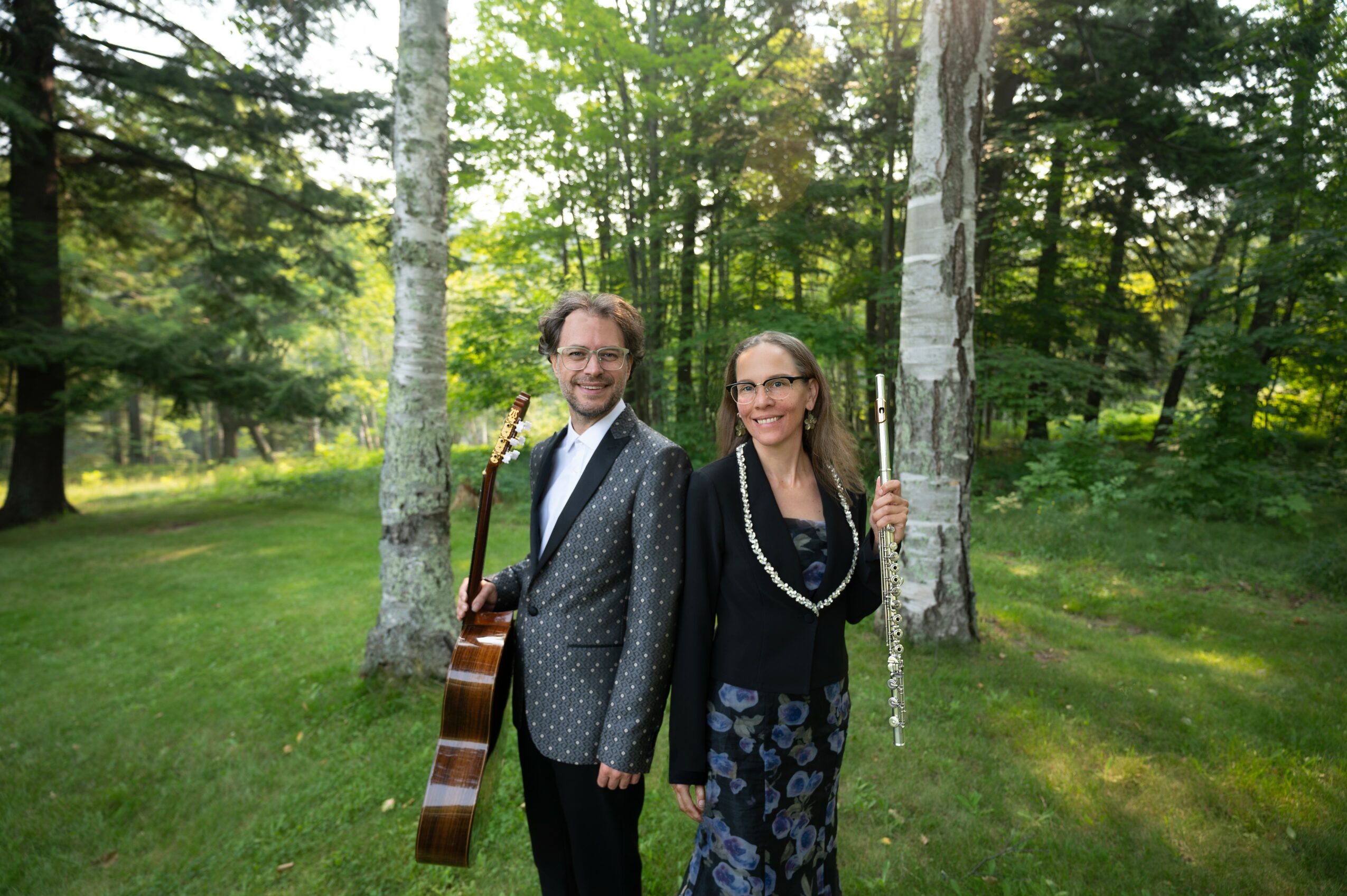 Two people stand outdoors in a green park with trees, smiling, one holding a guitar, the other holding a tree trunk.